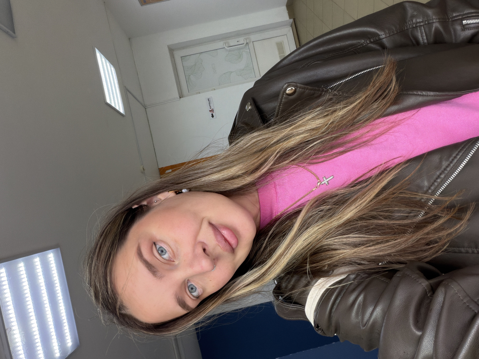 The image shows a woman with blue eyes, light brown hair, wearing a pink top and a brown leather jacket. She is in an indoor setting with a bright LED light fixture on the wall. A white door and some wall-mounted equipment are visible in the background. Based on the available information, it is impossible to confirm the exact location in Chaikovsky, Russia, without further context.