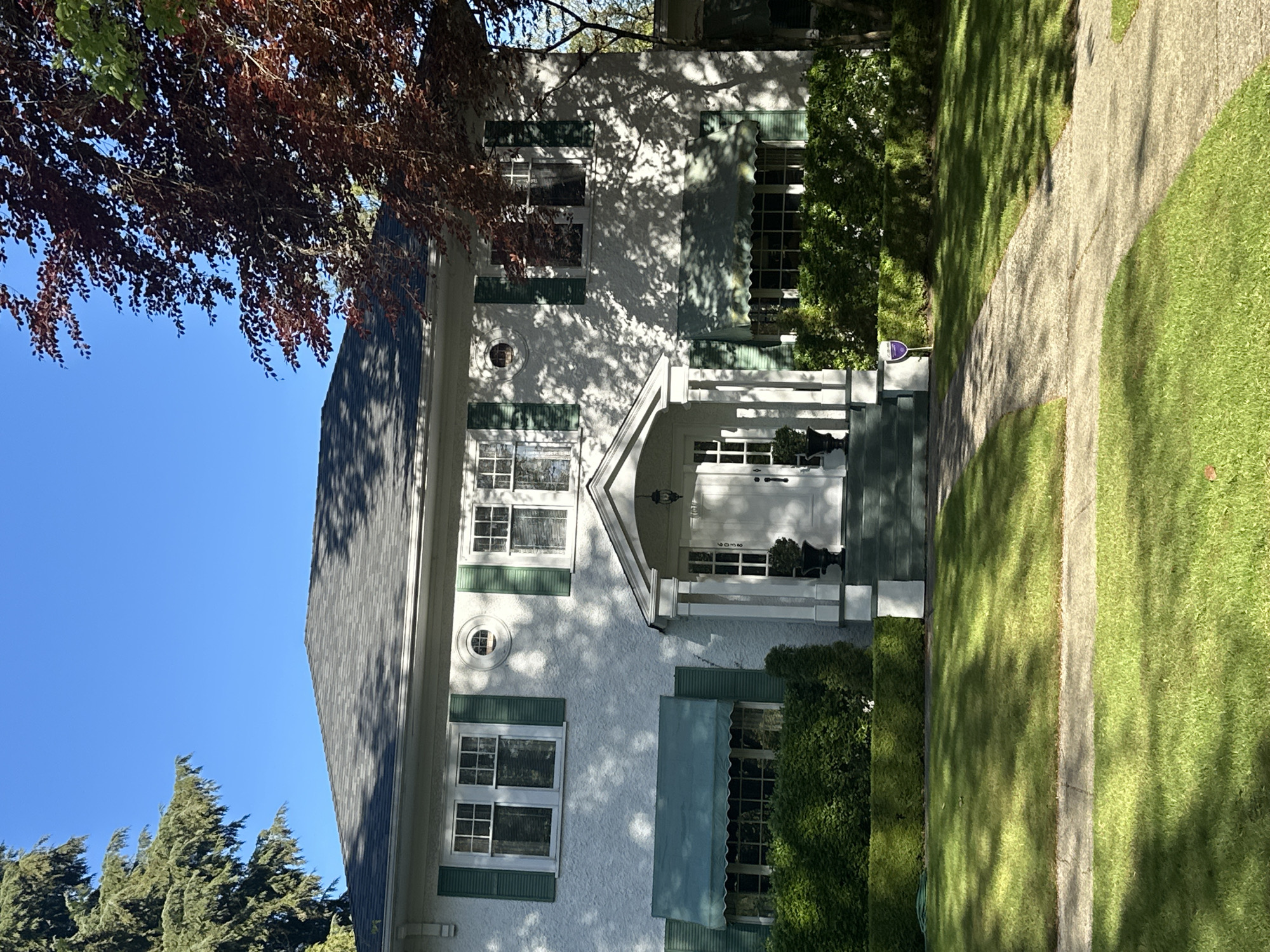 The image, rotated 90 degrees counter-clockwise, depicts a two-story residential house in bright daylight, likely in Vancouver, Canada. The scene captures the front facade of the house, a manicured lawn, a winding pathway, and surrounding vegetation.
The primary subject is a well-maintained house with a light-colored, possibly stucco, exterior and a dark shingled roof. It features multiple windows with white frames and dark green shutters, adding a traditional aesthetic. Several windows on the ground floor and upper floor are adorned with dark green fabric awnings. A prominent white portico with pillars frames the main entrance, above which the house number "2052" is faintly visible. A small, round window is also present on the upper facade. Strong shadows from overhead trees are cast across the house's exterior, indicating direct sunlight.
To the right of the house (when viewed upright), a light-colored, curved pathway meanders through a lush green lawn. The lawn itself shows dappled shadows from trees, suggesting a sunny day with clear skies. Dense green hedges line parts of the property, providing privacy and landscaping. In the foreground, a large tree with dark reddish-brown leaves hangs down from the top left. In the background to the left, tall evergreen trees with needle-like foliage are visible against a clear blue sky.
No people or explicit activities are visible, presenting a peaceful, static street scene. A small purple device or sign is visible near the front steps, but any text on it is too blurry to discern. The overall impression is one of a well-kept home on a pleasant, sunny day.