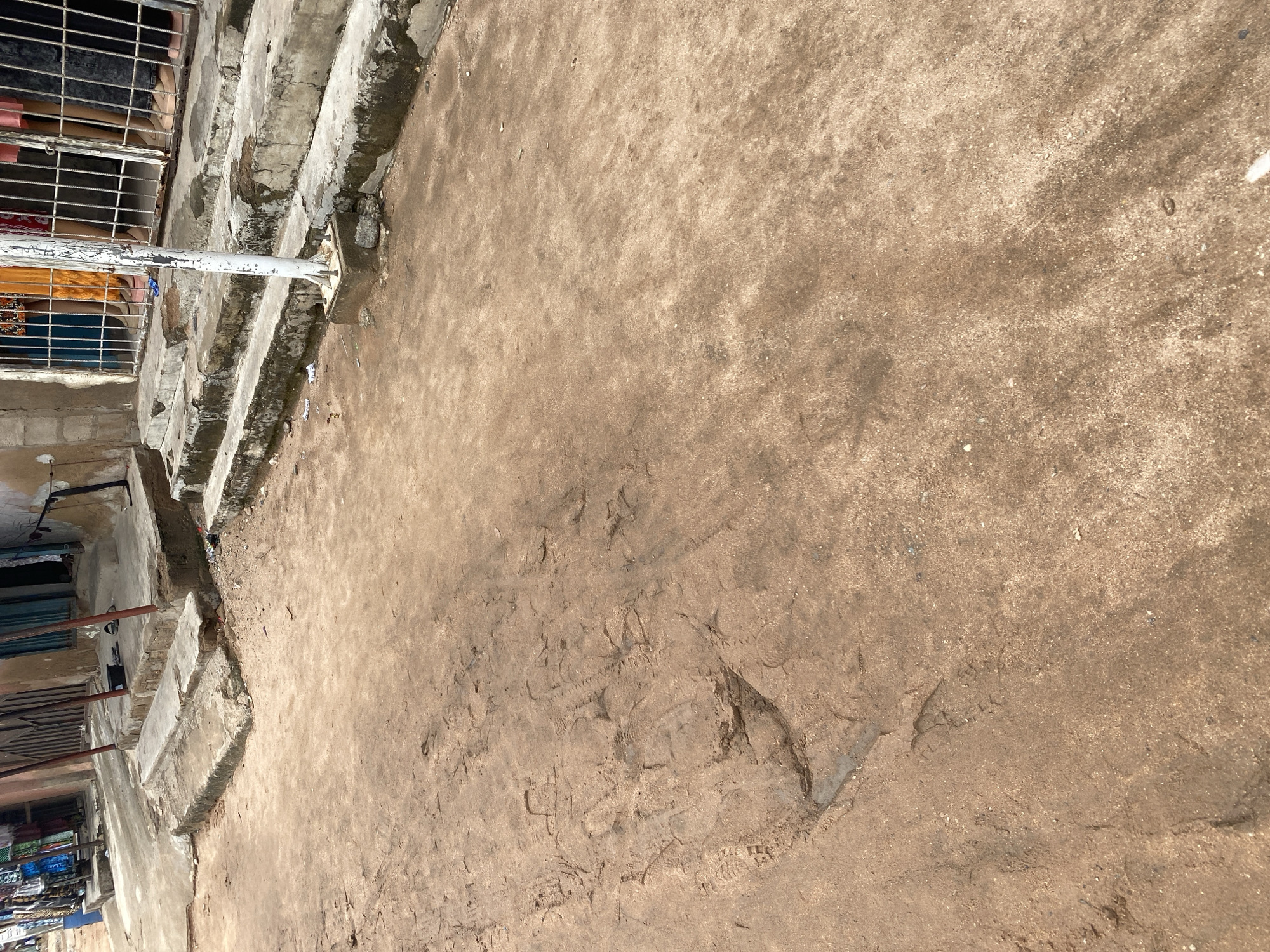 The image depicts an outdoor scene, likely a marketplace or a street in Jalingo, Nigeria. In the foreground, a dusty, uneven ground is visible, marked with tire tracks and footprints, suggesting recent activity. To the left, a series of concrete steps leads up to a building with metal bars, behind which are glimpses of people and what appears to be merchandise, possibly textiles or clothing. A white pole, likely part of a structure or fence, stands prominently in the middle ground, dividing the scene. The overall impression is one of a bustling, everyday environment. The lighting suggests it is daytime, possibly mid-morning or afternoon, with no immediate indicators of specific weather conditions. The context of Jalingo, Nigeria, places the scene within a West African urban setting. No visible text is present in the image.