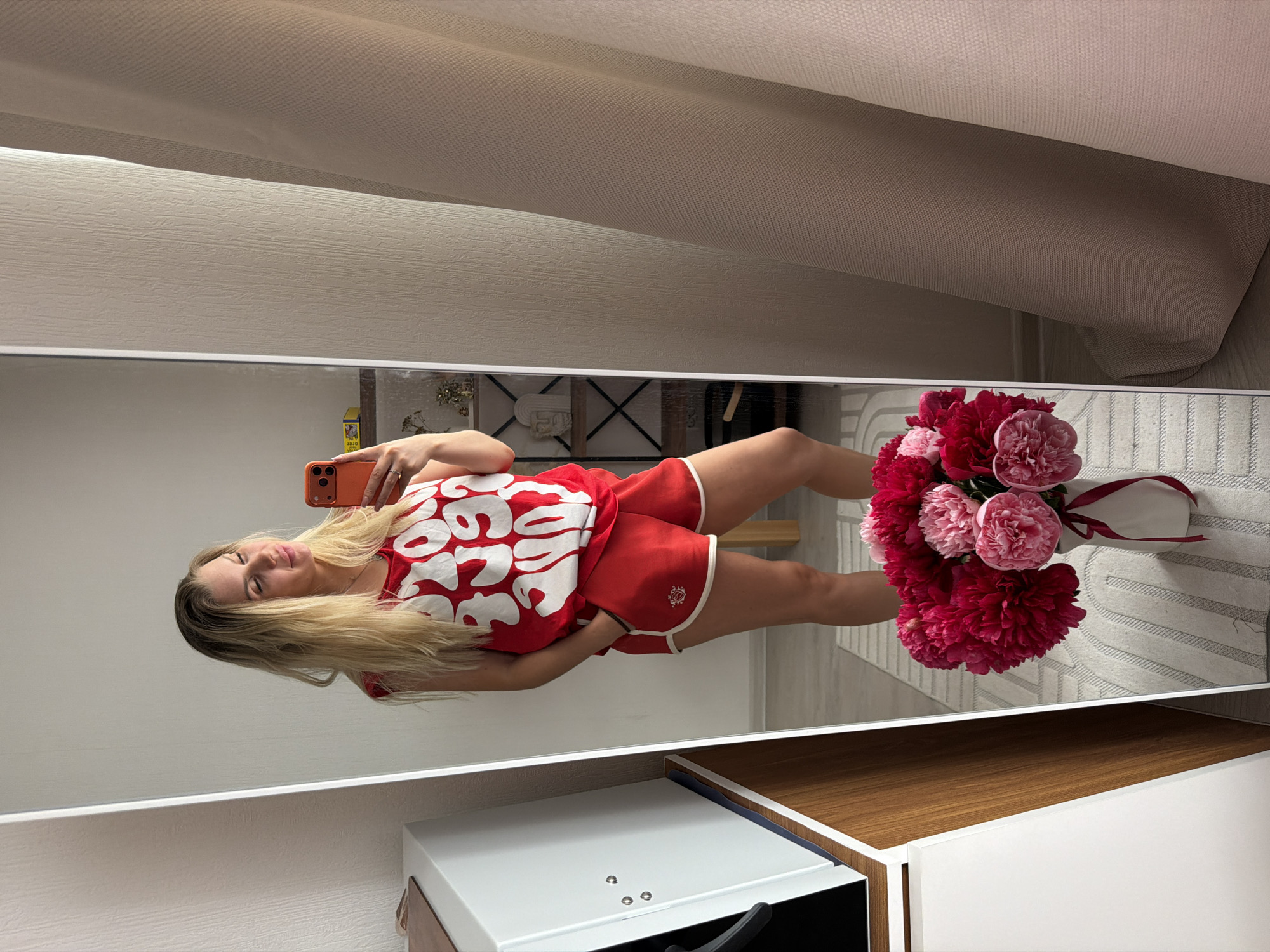 A woman with blonde hair stands in front of a full-length mirror, taking a selfie with an orange smartphone. She is wearing a red tank top with white lettering and red shorts with white trim. Her hands are in her pockets. In the foreground, reflected in the mirror, is a bouquet of pink and red peonies in a white vase tied with a red ribbon. The room appears to be a bedroom or living area, with a white cabinet to the left, shelves with decorative items in the background, and beige curtains to the right. The lighting is soft and natural. The location is not specified as Moscow, Russia, and there is no information in the image to support this claim.