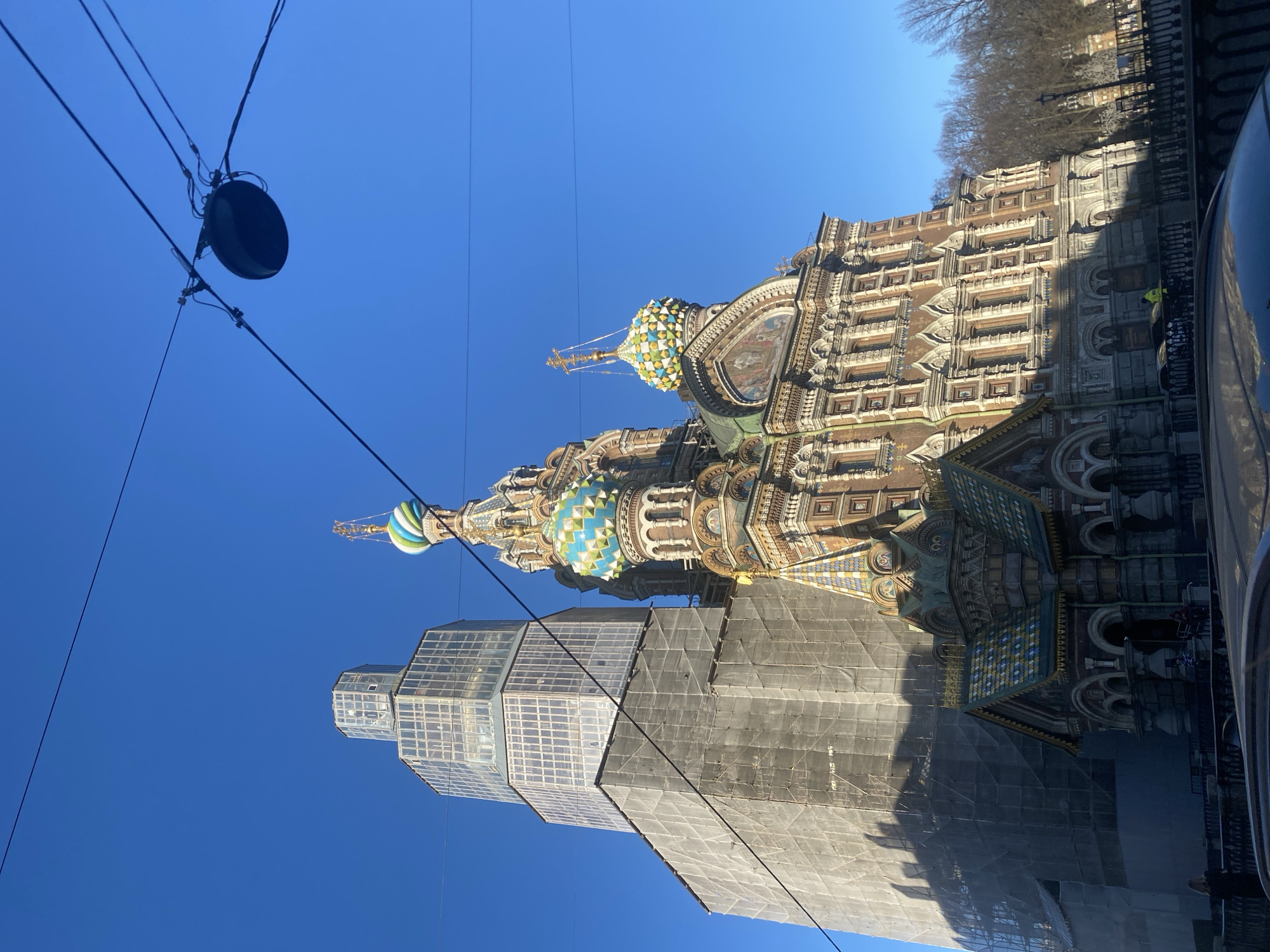 The image shows the Church of the Savior on Spilled Blood in Saint Petersburg, Russia. The church's colorful onion domes and ornate facade are visible, and the building appears to be undergoing restoration, with scaffolding and protective coverings present on a portion of the exterior. Electrical wires and a streetlamp are visible in the foreground against a clear blue sky. Trees and a building are in the background, and the edge of a vehicle can be seen on the right side of the frame.