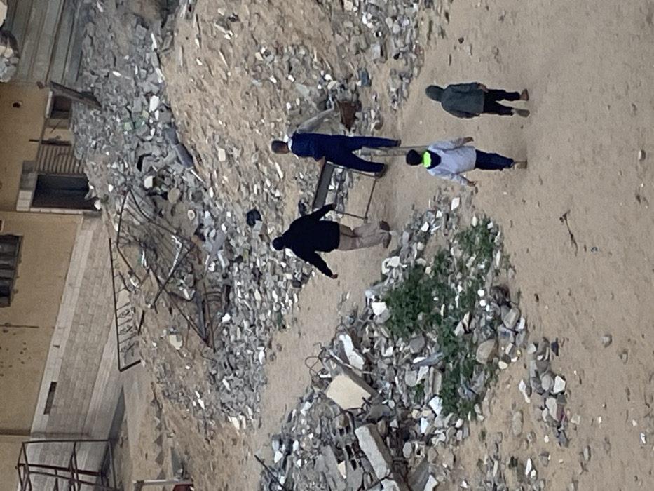 From a high angle, the scene depicts four people walking through a debris-strewn landscape. The debris, consisting of gray and white rubble, suggests a destroyed building. A damaged building's facade is on the left. The people are dressed in casual attire. A patch of green vegetation grows amidst the destruction. The ground is sandy or dusty. The overall impression is one of devastation.