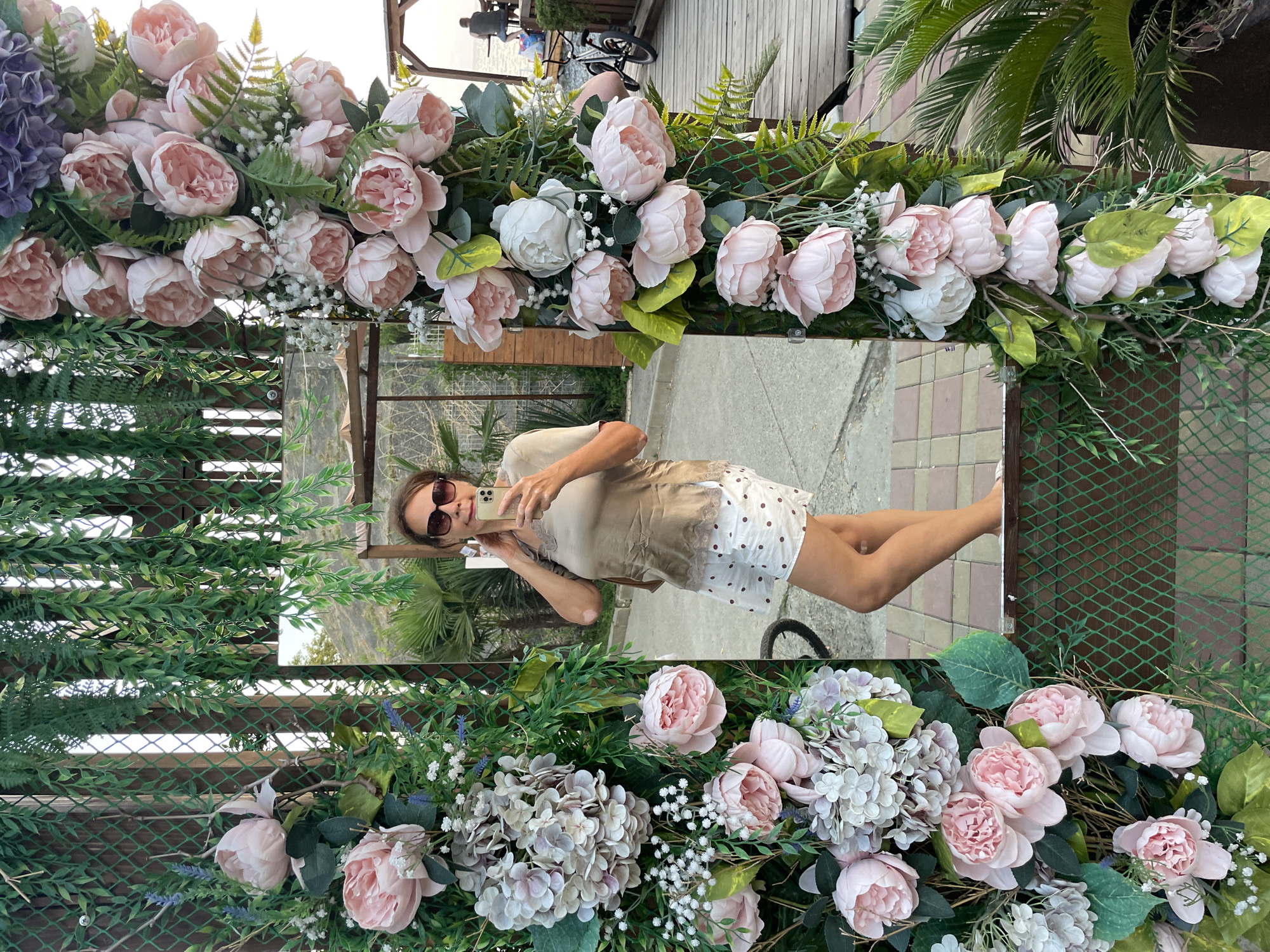 A woman is taking a selfie in a mirror, surrounded by floral decorations. She is wearing sunglasses, a beige top, and a white skirt with brown polka dots. The mirror is framed by artificial pink and white peonies and greenery. In the background, through the mirror, a man is sitting at a table on a wooden deck near the sea. The scene appears to be outdoors in a public area, possibly a cafe or restaurant, located in Sochi, Russia, given the palm trees and coastal scenery.