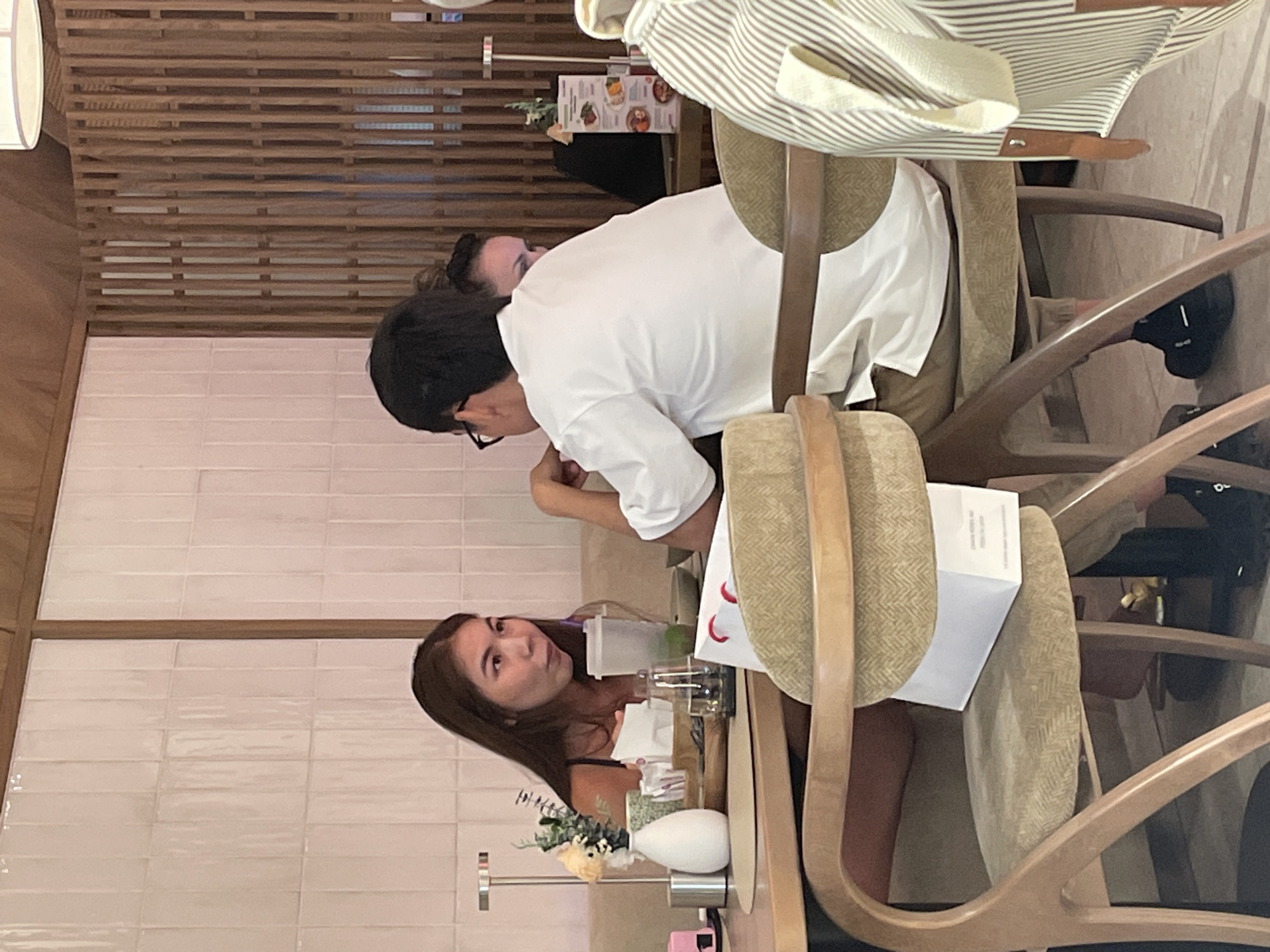 The image shows a group of people seated at tables in what appears to be a modern restaurant. In the foreground, a woman with long brown hair is looking to her right and speaking, with a drink in front of her. To her right, a man with dark hair and glasses is seated, facing away from the camera. Another person's face is partially visible between them. The restaurant has a contemporary design with light-colored tiled walls and wooden slatted partitions. On the table, there are various items including a white vase with flowers, napkins, drinks, and a white bag. The chairs are upholstered in a textured fabric. The overall atmosphere suggests a casual dining experience. The restaurant is located in Almaty, Kazakhstan.