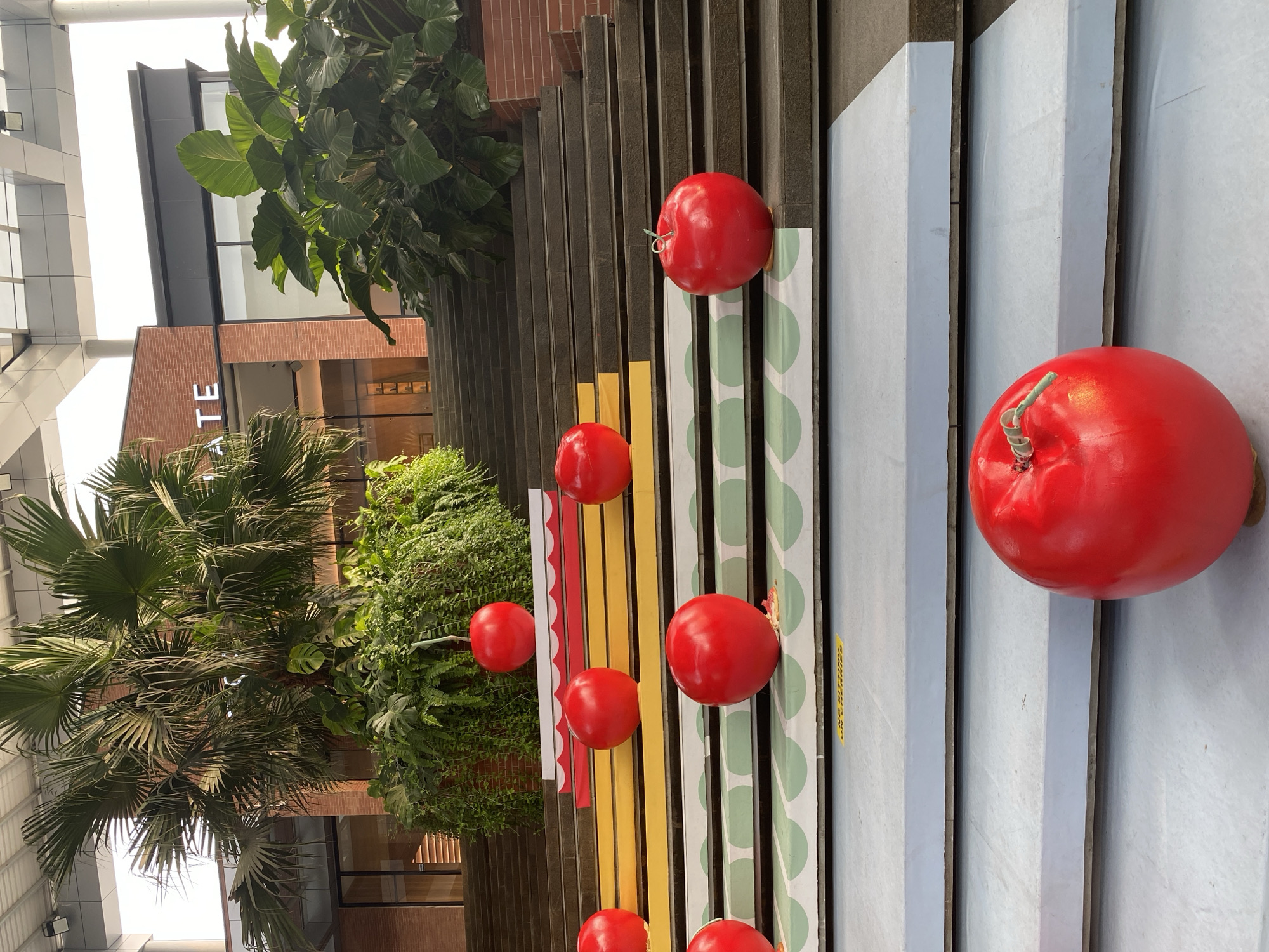 The image shows a festive outdoor staircase decorated with large red spheres resembling apples or cherries. The staircase is made of dark stone, with some steps adorned with colorful yellow, white, and light green striped patterns. Lush green plants, including palm trees and large-leafed foliage, frame the scene. In the background, a modern building with brick and glass facade is visible, with the word "GATE" displayed on the upper section. A hint of the location being in North Jakarta, Indonesia, is suggested by the context provided. The overall impression is of an art installation or decorative display designed to attract attention in a public space. There are no people visible in the image.