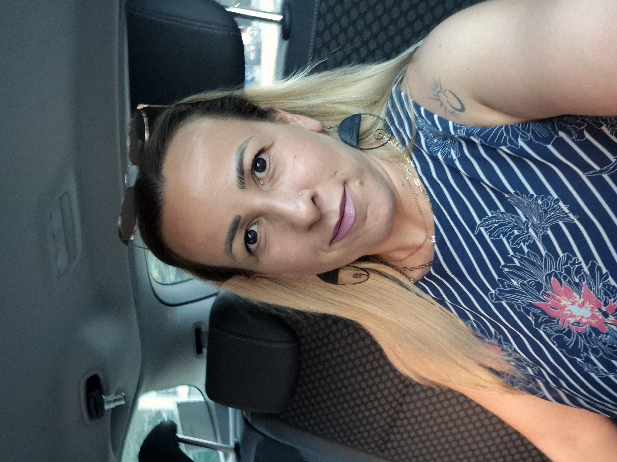 A woman with blonde hair and dark eyes, wearing a blue and white striped dress with a floral pattern, is sitting in the back seat of a car. She has a tattoo on her right shoulder. Her hair is partially obscured by the car's headrest. Sunglasses are perched on top of her head. The interior of the car is visible behind her, including the seat fabric and headrests.