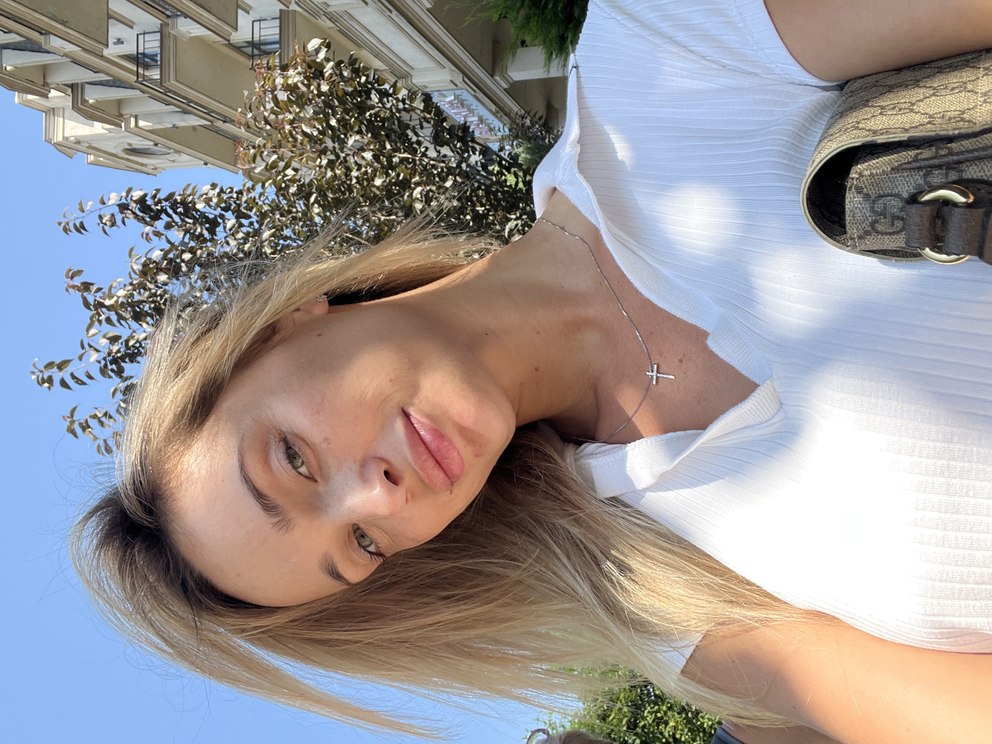 A blonde woman wearing a white ribbed shirt and a silver cross necklace is holding a brown designer handbag. She is outdoors, with a building and trees in the background. The sky is clear and blue.