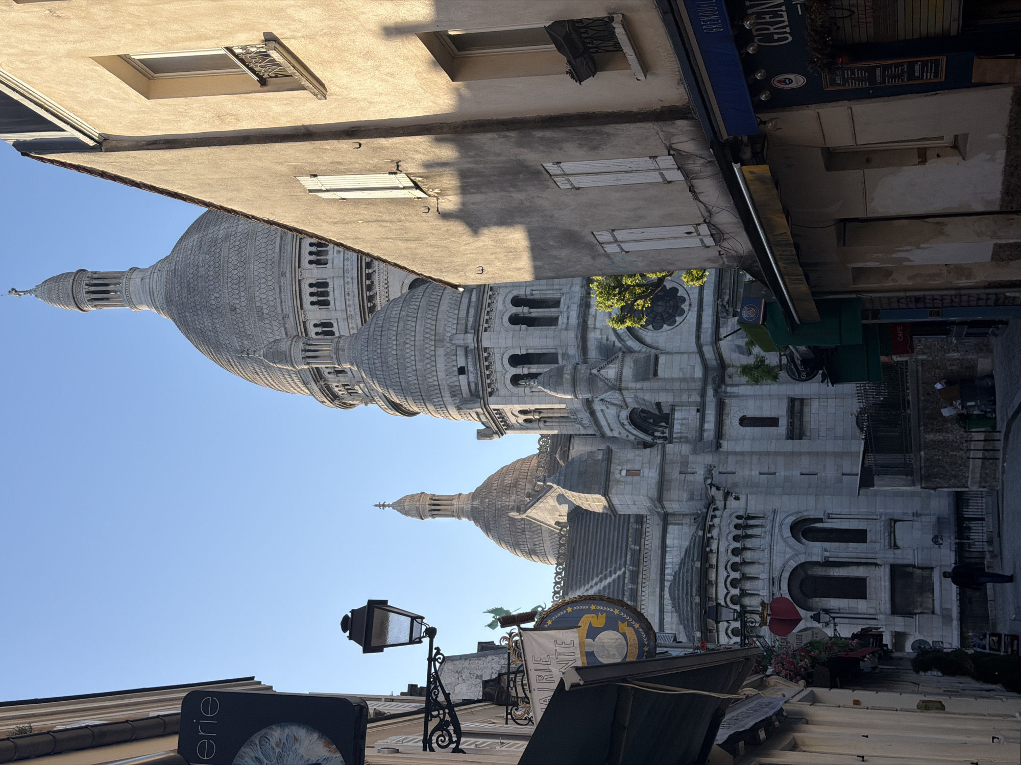 The image depicts a street scene in Paris, France, with the Sacré-Cœur Basilica prominently featured. The basilica's domes and architecture dominate the view. The street is lined with buildings, including a cafe with a blue awning labeled "Grenouille." Street signs and lamp posts are visible. A person is seen walking in the background along the street. The sky is clear and blue.