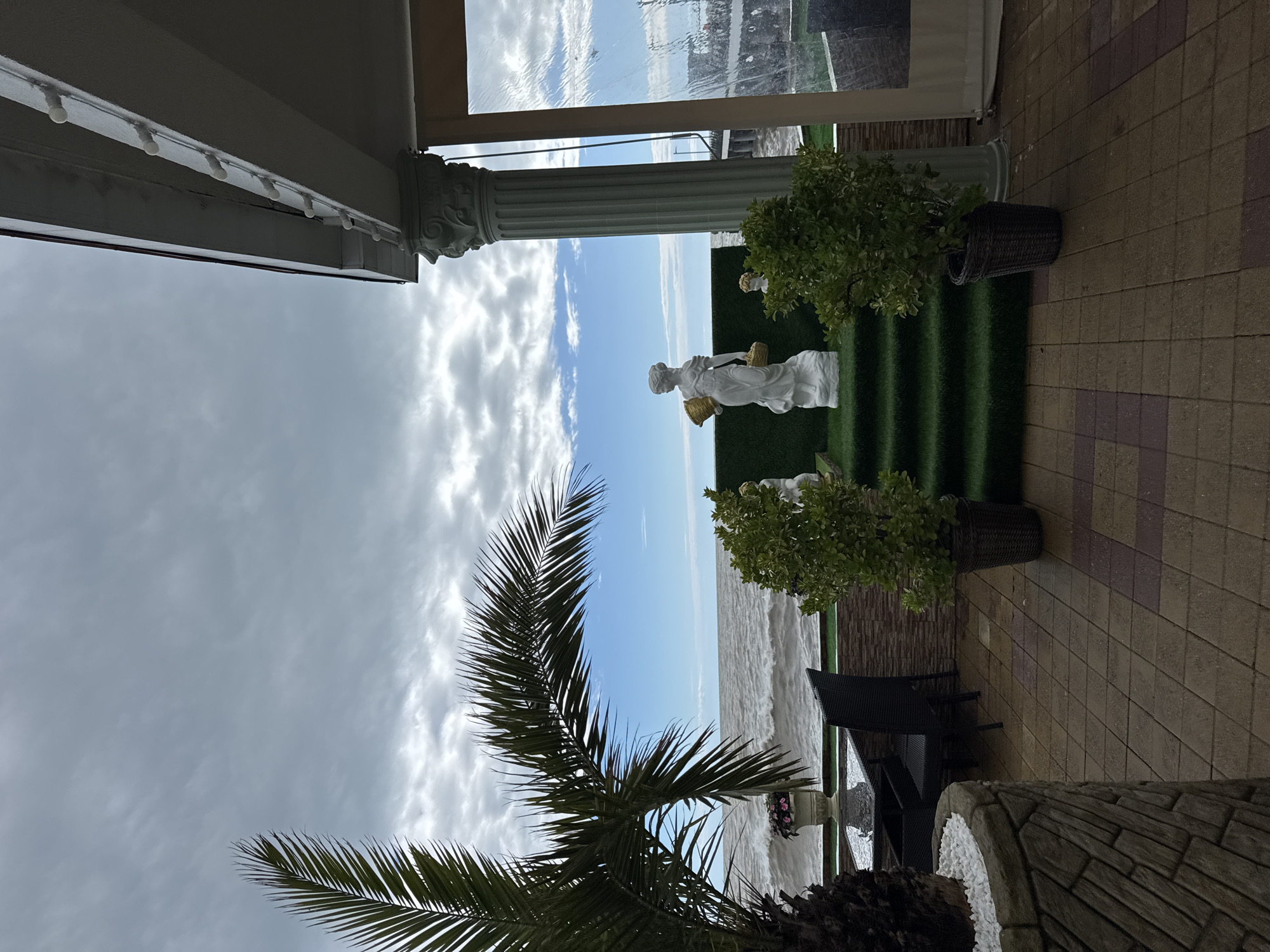 The image shows an outdoor terrace, possibly in Sochi, Russia, given the coastal view and architectural style. A white sculpture of a woman is the focal point, placed on a tiered, green artificial turf platform. The sculpture is flanked by potted plants. In the background, the sea meets the sky with a visible horizon line. The sky is partly cloudy. A palm tree is visible in the foreground. The terrace is paved with light brown and purple brickwork. Wicker furniture sits on the patio. A classical column supports a roof structure on the left side of the image.