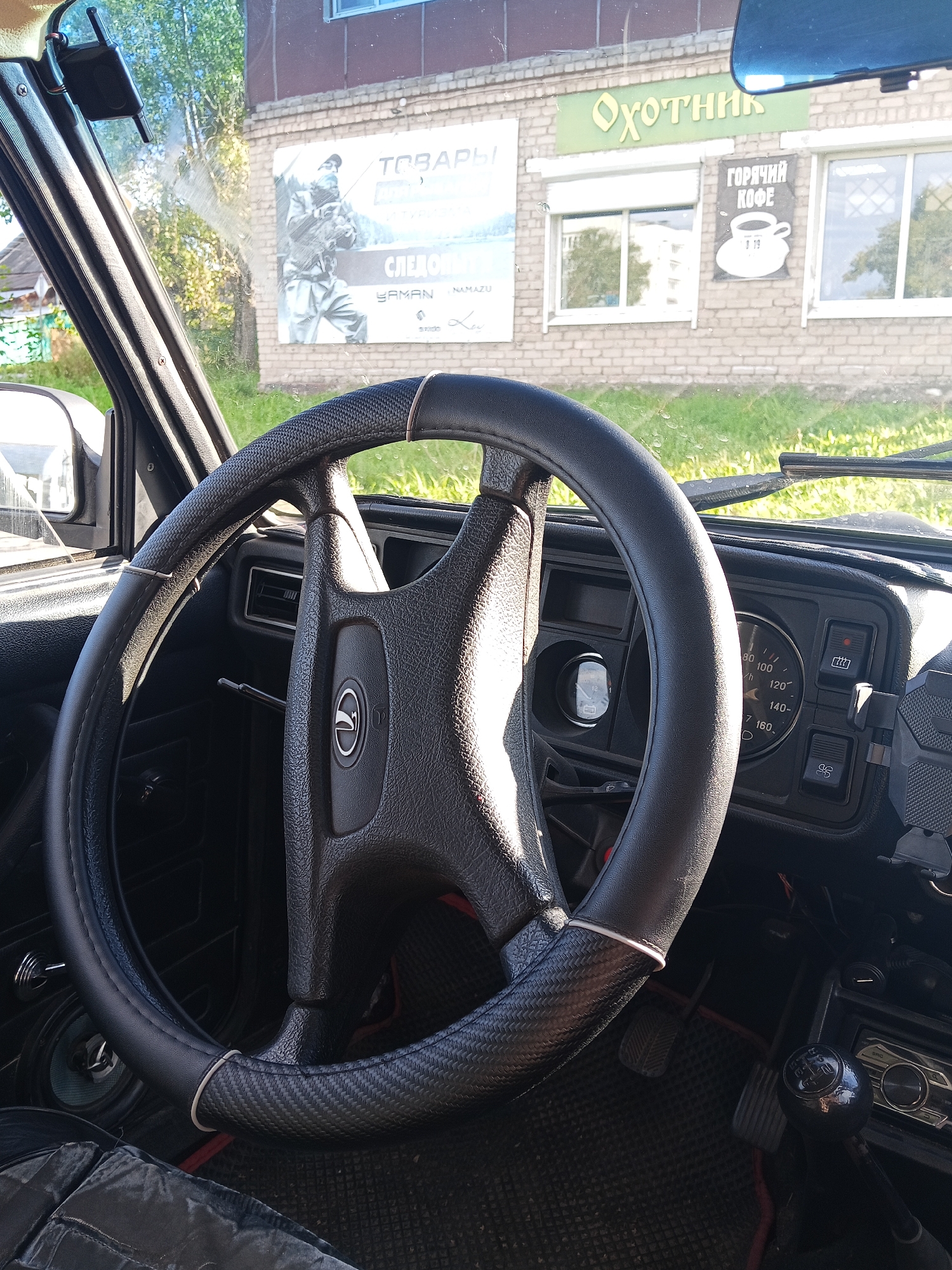 This is a medium shot, taken from inside a car, looking out the windshield at a building with a sign that reads "ОХОТНИК" (Hunter). The car's steering wheel and dashboard are prominently featured in the foreground. The dashboard includes a speedometer, fuel gauge, and various switches. The building appears to be a storefront, with a window displaying a sign for "ГОРЯЧИЙ КОФЕ" (Hot Coffee). The exterior of the building is brick, and there are some trees and grass visible outside. The overall scene suggests a stop at a roadside establishment in Chaikovsky, Russia.