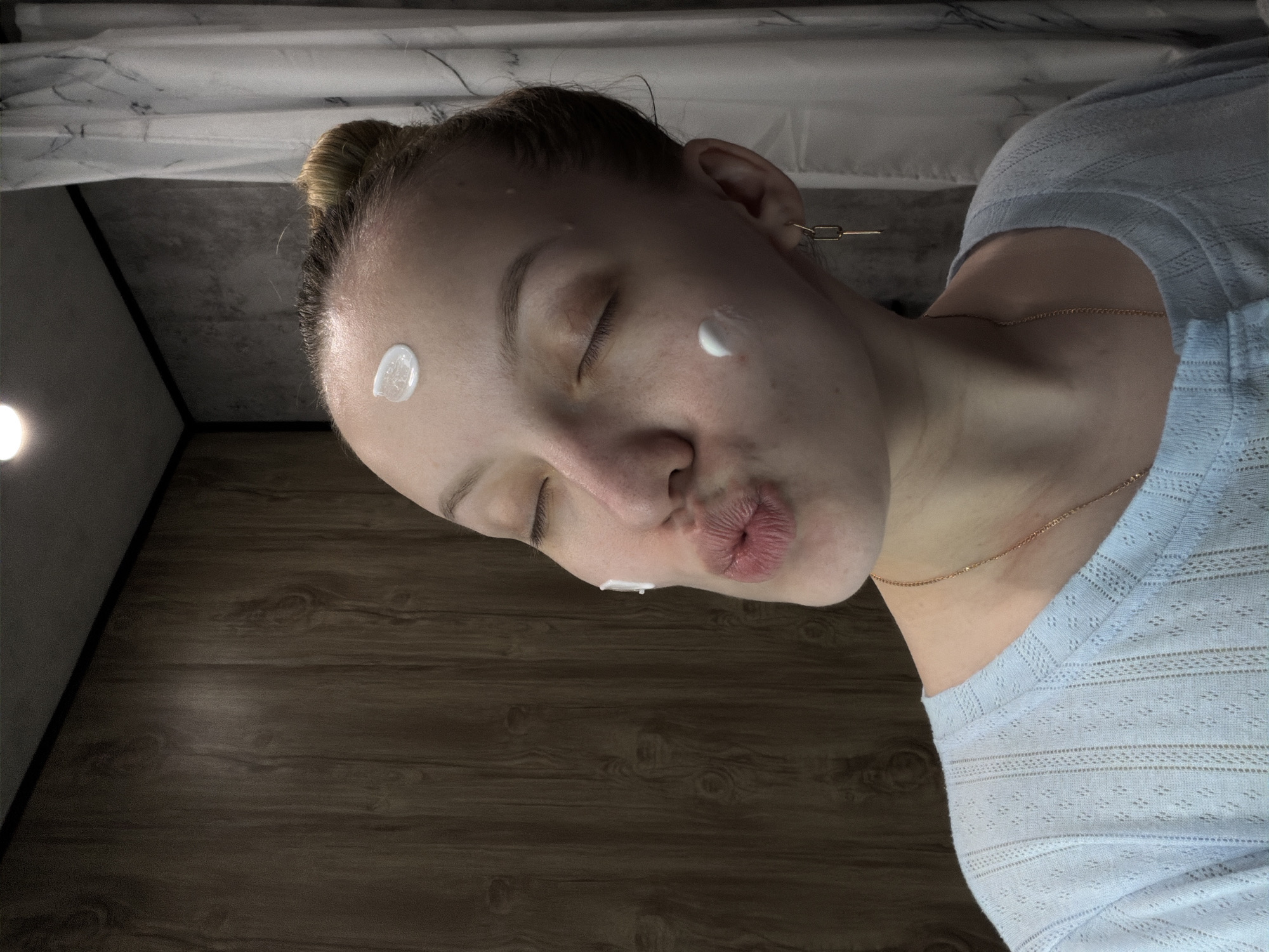 A young woman with fair skin and her hair in a bun poses for a selfie. She has white cream or lotion dotted on her forehead and cheek and is puckering her lips. She wears a light blue knit shirt and a gold chain necklace, and a gold chain earring is visible. The background shows a white marble-patterned surface above and a dark wood-grain surface below, suggesting a bathroom setting. This image does not contain sufficient information to place the subject in Reshetnikovo, Russia.