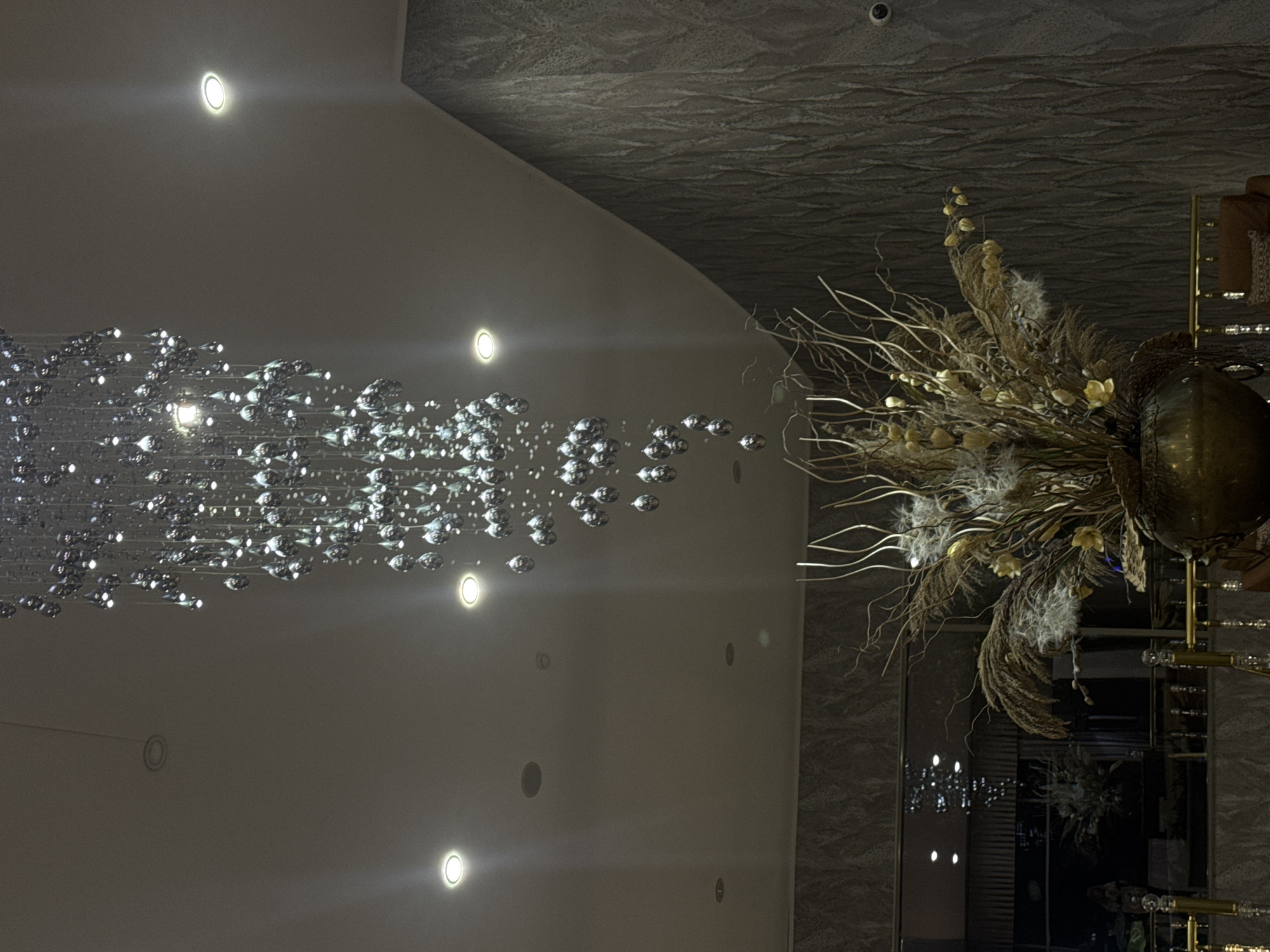The image displays an interior architectural scene, captured at an oblique angle, primarily focusing on ceiling and wall decor. The composition is horizontally oriented, but the visual elements suggest a potential vertical perspective if the image were rotated.
On the left, a large, ornate chandelier extends diagonally, appearing to cascade downwards from the upper left. It is composed of numerous small, sparkling crystal or glass elements, likely teardrop or spherical in shape, suspended on thin wires. These elements intensely reflect light, creating a shimmering, flowing effect across a smooth, light grey ceiling. Several circular, recessed spotlights are also embedded in this ceiling section, emitting bright, focused beams.
The right side of the image features a darker, textured ceiling or wall surface with a distinct wavy or rippled pattern, meeting the lighter ceiling section at an angle. A small security camera (CCTV) is discreetly visible on this textured surface near the top edge. Below this, a large, striking decorative arrangement made of dried natural elements, including slender branches, feathery pampas grass, and other dried flora, is prominently displayed. This organic centerpiece offers an earthy, bohemian contrast to the modern chandelier. It is mounted on or stands before a dark, reflective panel, possibly a mirror, which shows blurred reflections of interior lights and other elements.
The scene conveys a sense of sophisticated interior design, characteristic of a high-end establishment such as a hotel lobby, a chic restaurant, or an upscale event venue. No people or activities are visible, and the primary reliance on artificial lighting suggests an indoor setting, possibly during evening hours. There is no discernible text within the image.