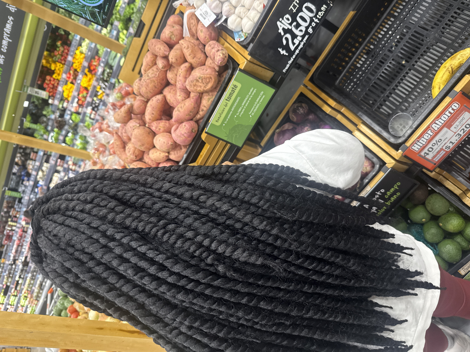 In Mosquera, Colombia, a person with long, dark, braided hair stands in a grocery store aisle. They are facing away from the camera, looking at a display of produce. The foreground is dominated by the person's elaborate braids. Behind them, shelves are stocked with various fruits and vegetables, including sweet potatoes, garlic, and avocados. Signage is visible, indicating prices and product information. The lighting appears to be typical of a supermarket environment.