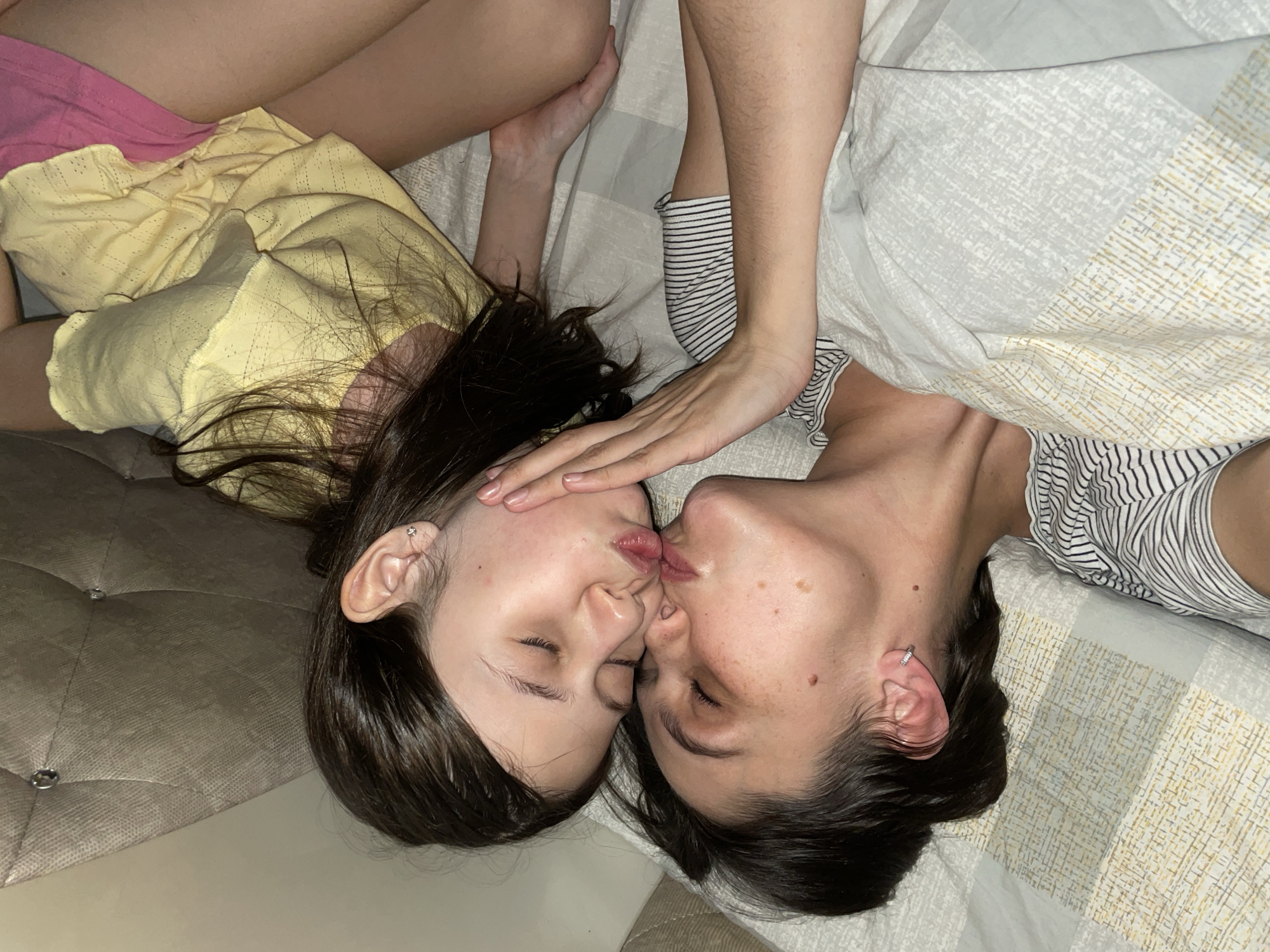 Two individuals are lying on a bed with patterned sheets and a quilted headboard. One is wearing a yellow top and pink shorts, while the other is wearing a striped top. They are kissing. The location is not discernible as Izhevsk, Russia, based solely on visual information from the image.