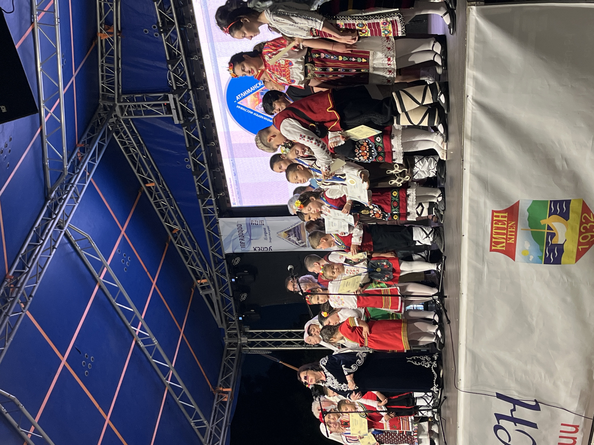 On a stage, a group of children and adults, most of whom are wearing traditional Bulgarian folk costumes, are gathered. Some of the children are receiving awards or certificates. The stage is set up outdoors, indicated by the truss structure supporting a blue tarp overhead and the dark night sky visible in the background. A large screen is positioned behind the group, displaying text and graphics. A banner with the text "КИТЕН" (Kiten) and the year "1932" is visible at the bottom of the image, suggesting the event is taking place in or related to the city of Kiten, Bulgaria. The overall scene depicts an awards ceremony or a cultural performance.
