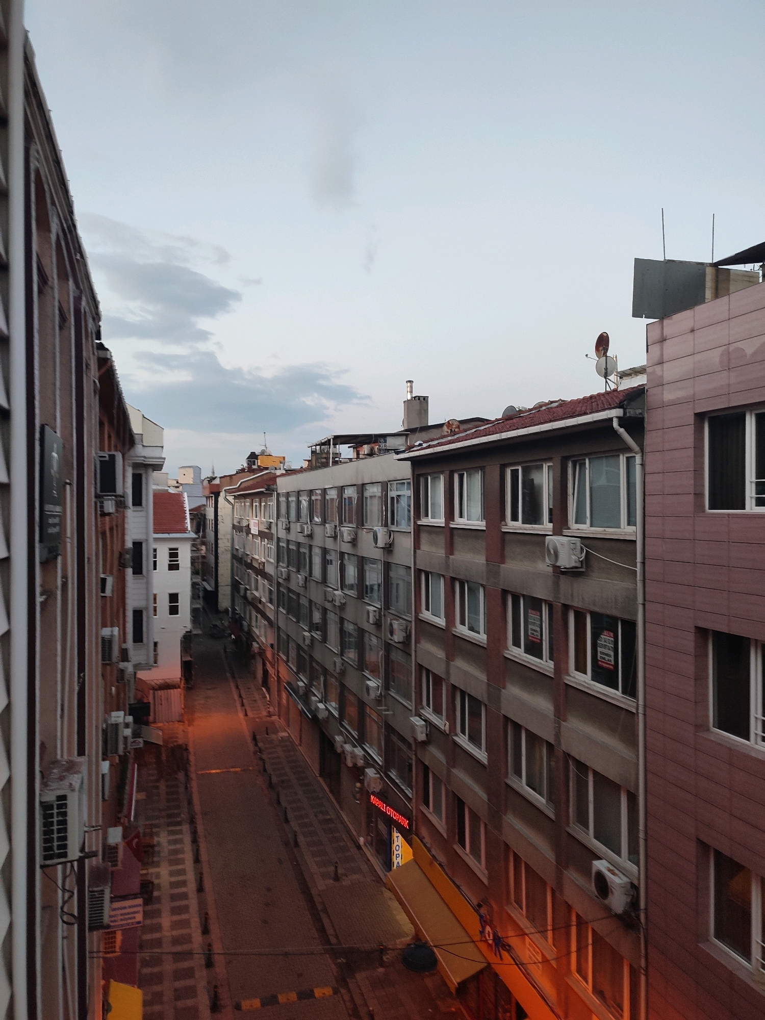 The image is a high-angle view of a narrow street in Kadıköy, Turkey, likely captured during dusk or twilight. The street is flanked by multi-story buildings, many of which have air conditioning units mounted on their facades. The buildings display a mix of architectural styles, with some featuring brick or tiled exteriors. Several windows have signs posted on them. A few streetlights cast an orange glow on the cobblestone street and sidewalk. A red neon sign that reads "KIZIL TOPRAK OTOPARK" is visible on the right side of the street. The sky is overcast with a pale grey hue. The perspective suggests the photo was taken from a balcony or window of a building overlooking the street. There are no discernible people in the image, and the scene appears to be quiet and mostly deserted.
