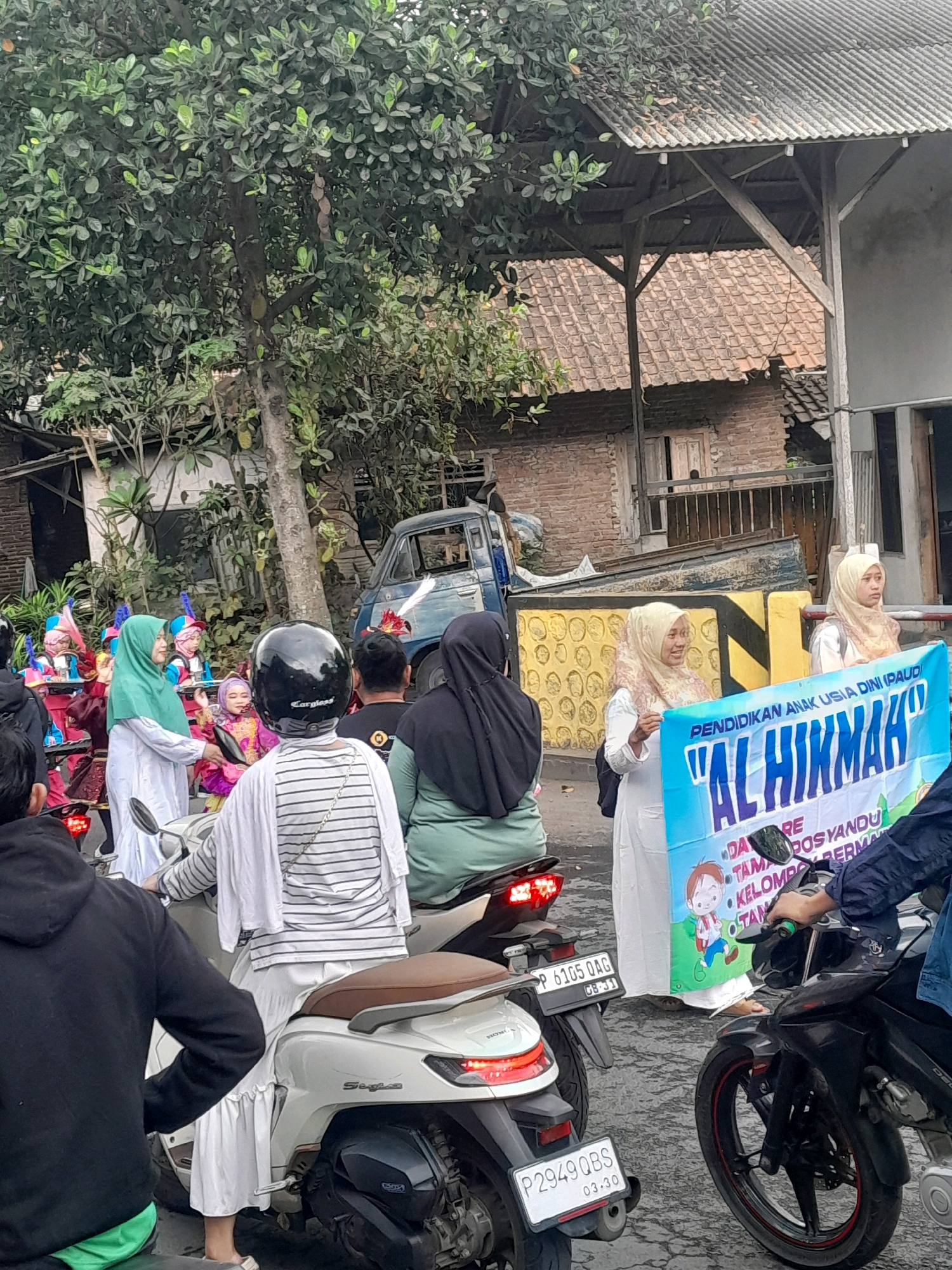 The image depicts an outdoor scene in Gentengwetan, Indonesia, likely during a parade or procession. In the foreground, several motorcycles are visible. One white scooter is ridden by a person wearing a black helmet and a white and grey striped top. Another black motorcycle is being ridden by someone wearing a dark blue jacket and jeans, holding onto the handlebars. The rear license plate of the white scooter reads "P 2949 QBS". The license plate of a motorcycle behind it reads "P 6105 OAG".
In the middle ground, a group of children dressed in colorful, festive attire, including headwear with feathers, are being led by adults. One adult, wearing a green hijab and white clothing, is holding a tray. Another adult, wearing a light-colored hijab and white clothing, is holding a banner. This banner, with blue and white lettering, reads "PENDIDIKAN ANAK USIA DINI IPAUD" and below that, "AL HIKMAH". There are illustrations of children on the banner.
In the background, a blue truck is parked. The environment includes buildings with tiled roofs and trees with green foliage. The ground appears to be paved. The overall impression is of a community event taking place on a street.