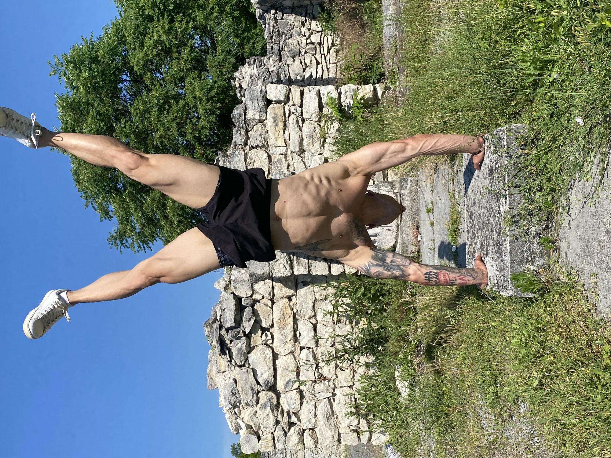 A shirtless man with tattoos on his arms and legs is performing a handstand outdoors. He is balancing on his hands, with his legs extended upwards in the air. He is wearing black shorts and white sneakers. The background consists of a stone wall and lush green vegetation under a clear blue sky. This scene is located at the Kaylaka Park in Pleven, Bulgaria, specifically in the ruins of the St. George's Chapel.