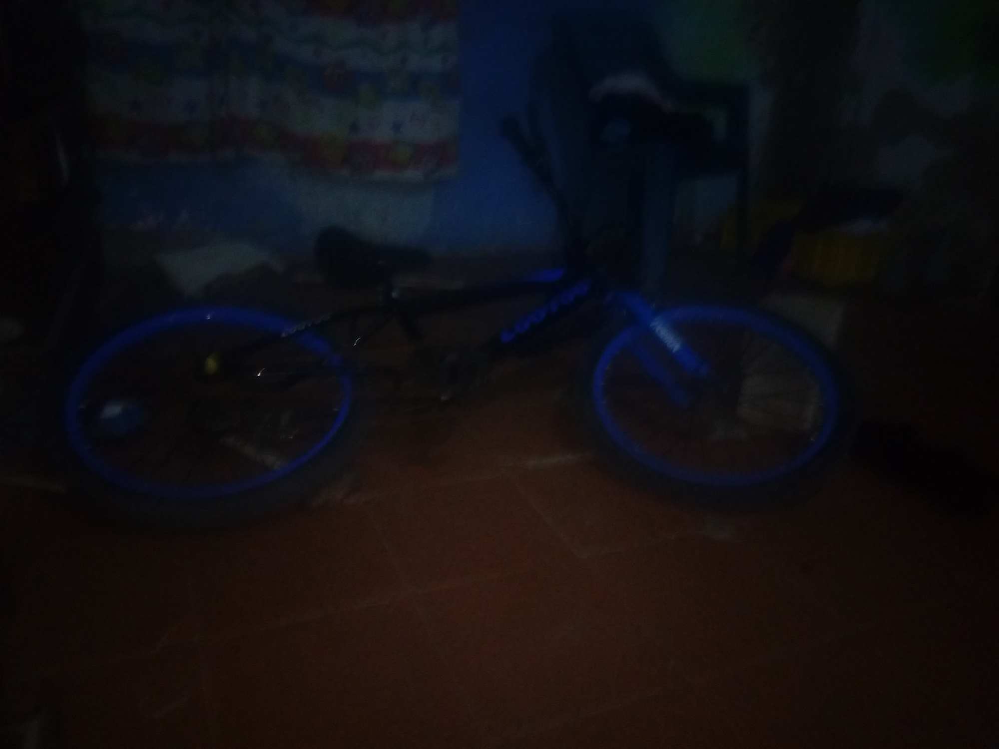 A black BMX bicycle with blue rims rests on a dark, reddish-brown tile floor. The bike is angled slightly to the right. In the background, there is a curtain with a colorful, patterned design. The scene is dimly lit, making details difficult to discern. The location is likely indoors in Juan Griego, Venezuela, based on request. No people are visible.