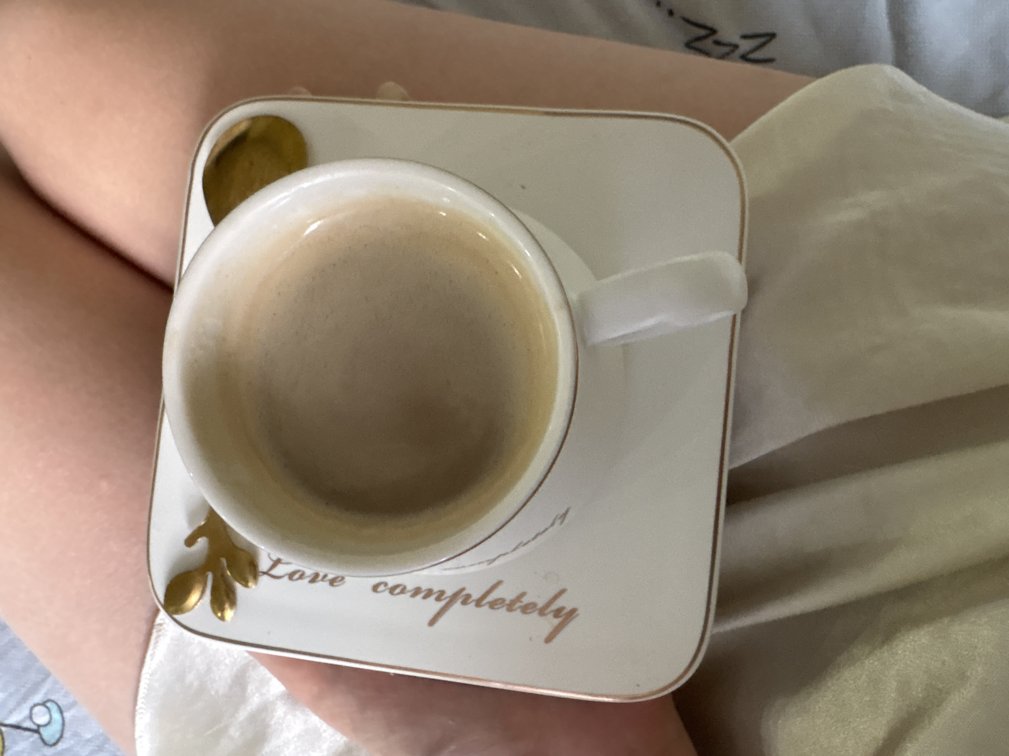 A high-angle close-up shot shows a cup of coffee resting on a saucer. The cup is white with a gold rim and a white handle. It is filled with a light brown coffee, potentially with milk. The saucer is square with rounded corners, adorned with gold decorations resembling leaves and the words "Love completely" inscribed in gold lettering. The cup and saucer are positioned on someone's lap, with a blurry background of skin and white bedsheets, partially revealing a bed with some patterned design. The setting appears to be casual and personal. The city of Chaikovsky, Russia cannot be confirmed from the image.