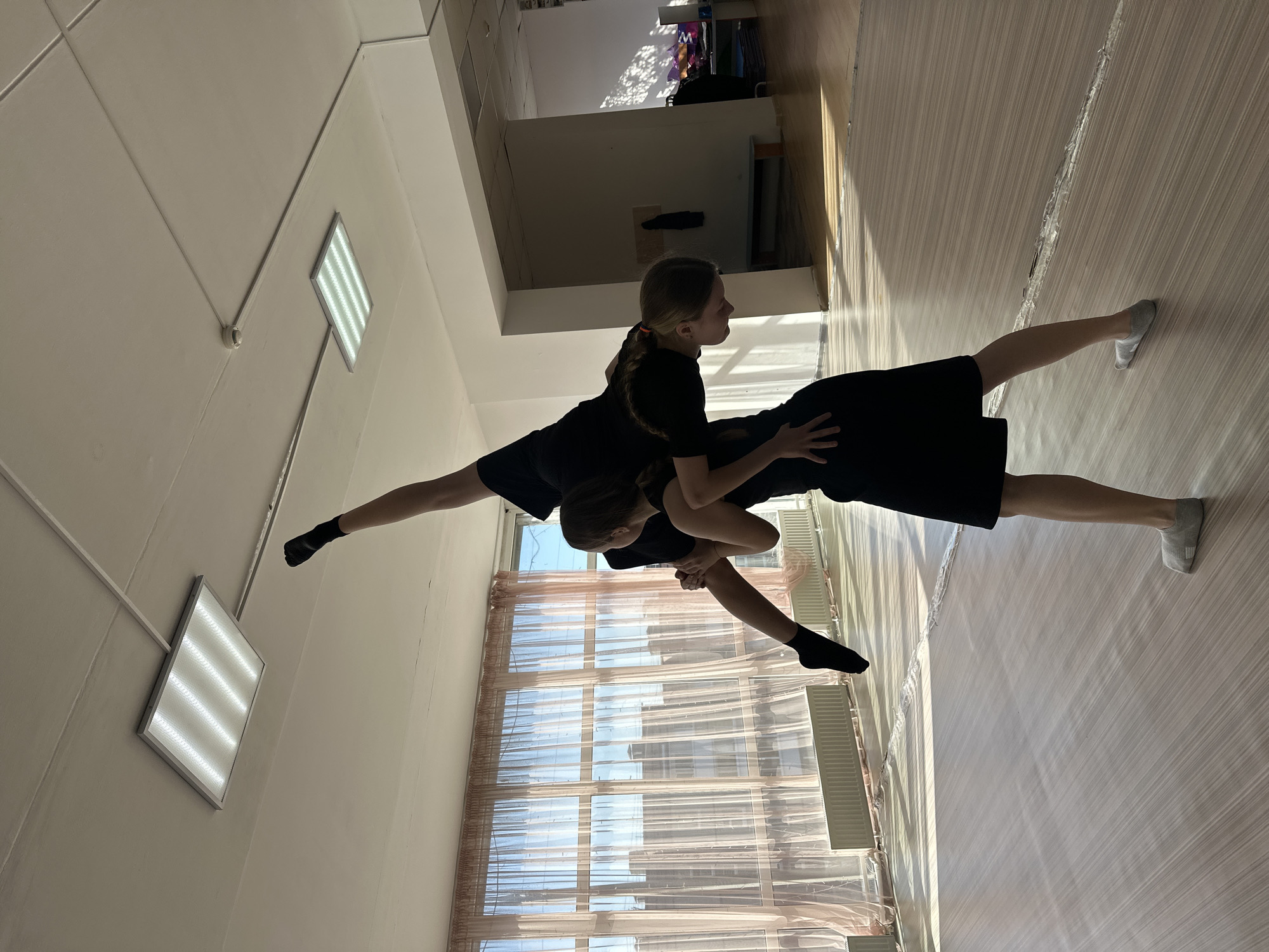 Two individuals are engaged in a dance pose within a brightly lit room. One person, with their legs extended outward, supports the other. The supported individual is positioned with their back towards the viewer, hands resting on the supporter's torso, head tilted slightly upward. Both are wearing dark clothing. The location appears to be a dance studio or similar space, evident by the light wooden floor, large windows covered with translucent curtains, and fluorescent lights on the ceiling. Through the windows, building exteriors and blue sky are visible. While precise identification of the location in Votkinsk, Russia, is not possible based on the image alone, the scene depicts a common dance practice environment.