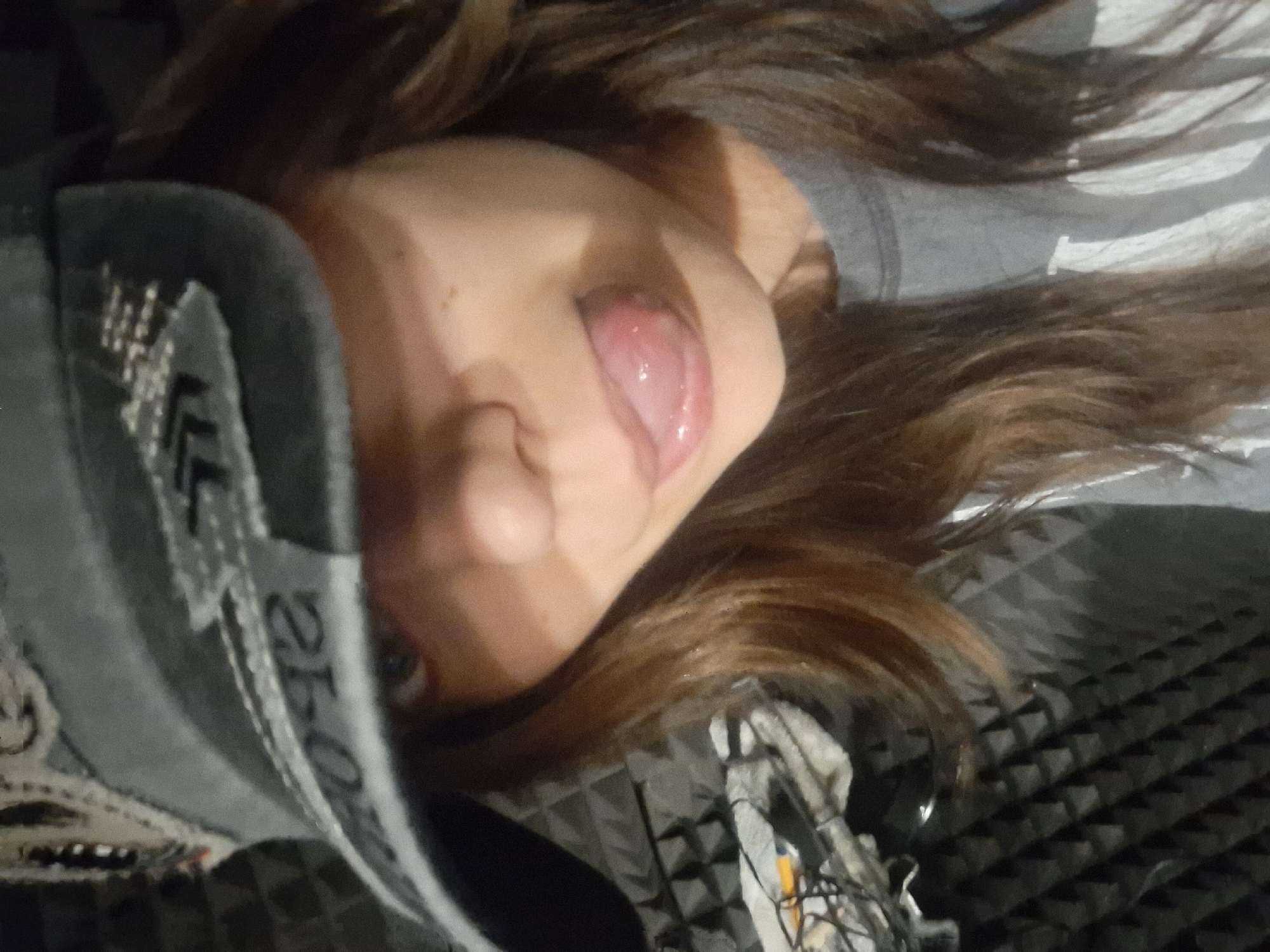 The image shows a person with brown hair sticking their tongue out. They are wearing a grey garment with white lettering, possibly a jacket or sweatshirt. The background features acoustic foam panels, suggesting an indoor recording or studio-like setting. There is no information to indicate the location in the city of Panagyurishte, Bulgaria.