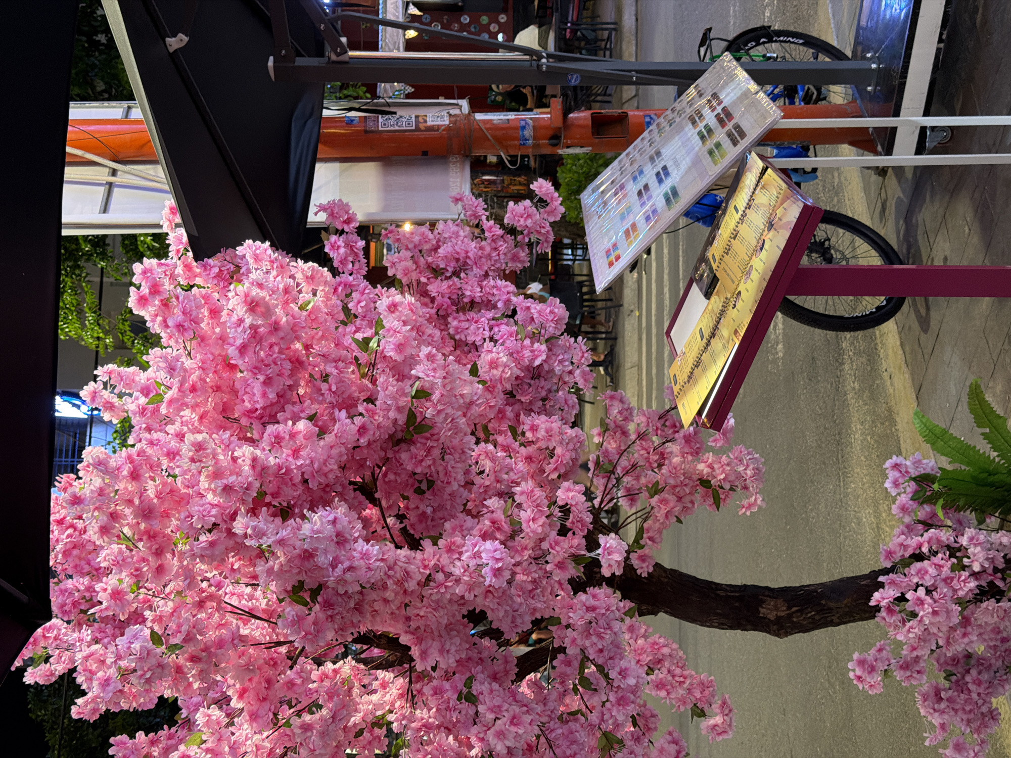 A pink artificial cherry blossom tree is in front of a restaurant with outdoor seating, displaying illuminated menu boards. A bicycle is behind the menus. The street is visible, but no people are clearly visible. This photograph may have been taken in the city of Đà Nẵng, Vietnam.
