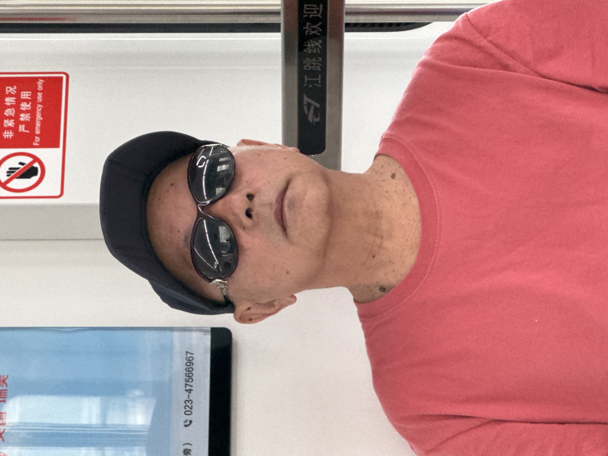 A medium shot captures a man inside what appears to be a Chinese subway or light rail car, with the image presented on its side. The man, positioned on the right side of the frame, is wearing a black baseball cap, dark sunglasses with silver frames, and a coral-colored t-shirt. He faces forward, looking towards the viewer, with his expression largely obscured by the sunglasses.
The setting is a clean, modern public transport vehicle with white interior walls. Behind the man, a vertical metal pole or screen displays Chinese characters that read "G 江跳线欢迎" (G Jiangtiao Line Welcome), indicating the vehicle belongs to the Jiangtiao Line. To the man's left, a red rectangular warning sign is visible, featuring a "no hand" icon. The sign displays text in both Chinese and English: "非紧急情况 严禁使用" (Non-emergency situation, strictly forbidden to use) and "For emergency use only."
Further to the left and partially visible is a blue vertical panel or advertisement. It shows a phone number, "023-47566967," where "023" is the area code for Chongqing, China, aligning with the Jiangtiao Line's location in Jiangjin, Chongqing. Fragmented Chinese characters like "圳天" and "旁)" are also visible on this panel. The image captures the man as a passenger, surrounded by the functional signage of the public transit system.