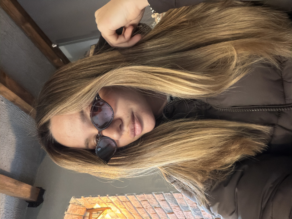 A headshot-style photograph depicts a single female individual indoors. The subject has long, straight to slightly wavy blonde hair with visible darker roots. She is wearing dark, angular-framed sunglasses with purple-tinted lenses and a dark brown puffer jacket, partially zipped, revealing a lighter-colored collar underneath. Her lips are slightly parted, and she is looking directly at the camera. Her left hand is raised towards her head, with fingers partially obscured by her hair, and a beaded bracelet is visible on her left wrist. The background to the left features a brick wall illuminated by a warm light source. Overhead, dark wooden beams are visible against a lighter-toned ceiling. The right side of the background is indistinct and darker. The scene is set in an indoor commercial or residential space in an unknown city.