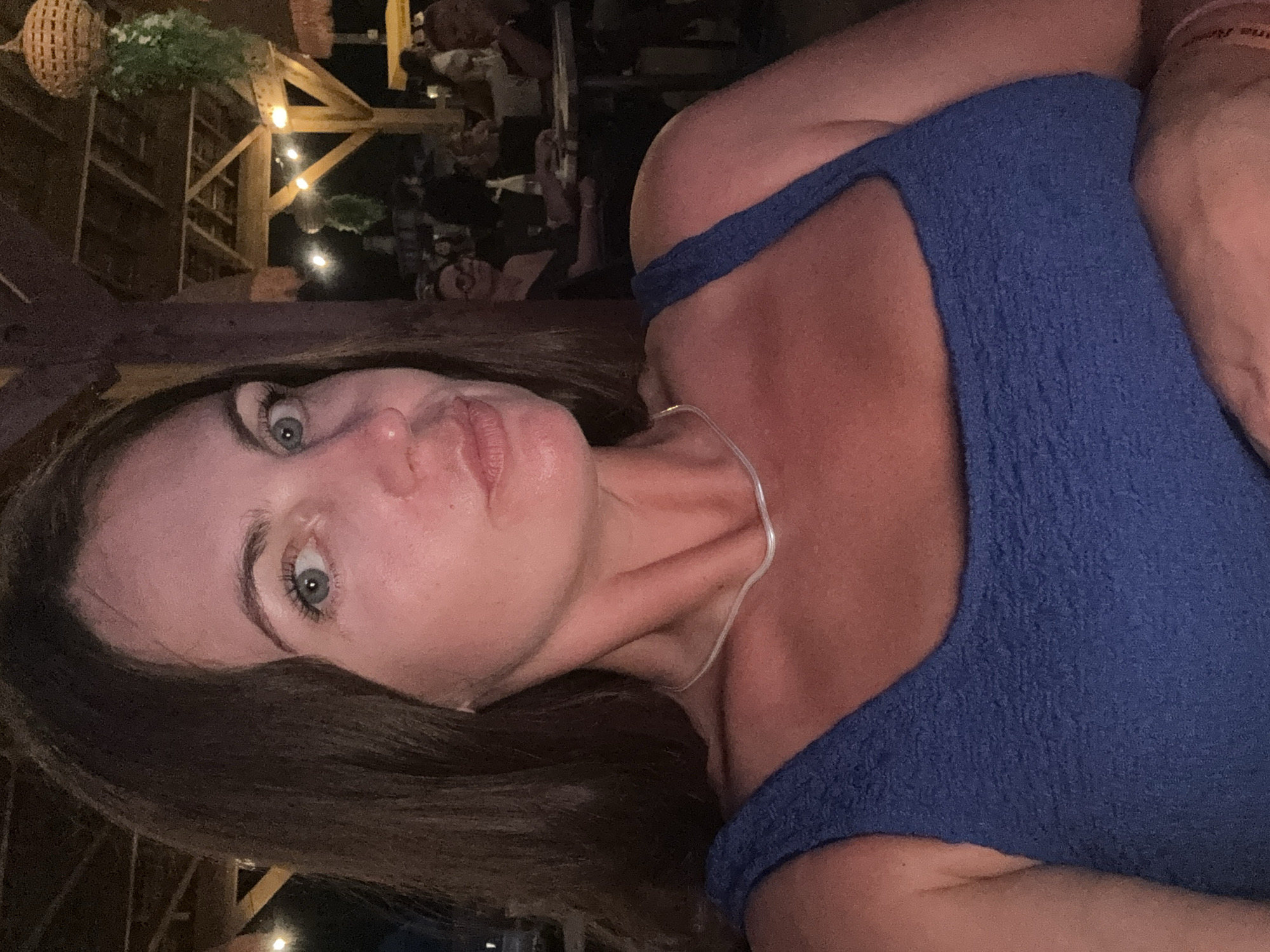 A medium close-up selfie shows a woman with medium-length brown hair and blue eyes looking directly at the camera. She is wearing a blue textured tank top and a thin silver necklace. The background appears to be an outdoor restaurant or bar setting at night, with visible wooden beams, string lights, and hanging greenery. Other patrons are blurred in the background, seated at tables. The image does not contain specific identifiable landmarks or characteristics that would definitively place it in La Grande-Motte, France.