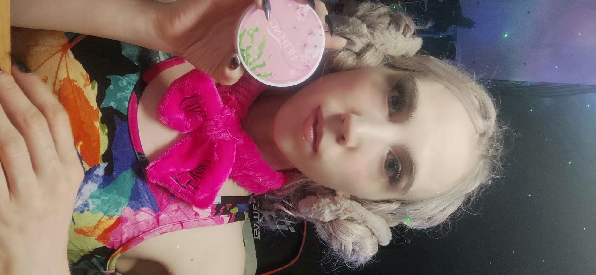 A woman with pale skin and platinum blonde hair styled in two buns is facing the camera. She is holding a round, pink compact with "LIFTHENG" written on it. She is wearing a brightly colored tank top with a floral pattern and a fluffy, pink scarf around her neck. The background includes a colorful desktop with a monitor displaying a starry night scene. The scene is indoors. There is no evidence suggesting the location is Chaikovsky, Russia.