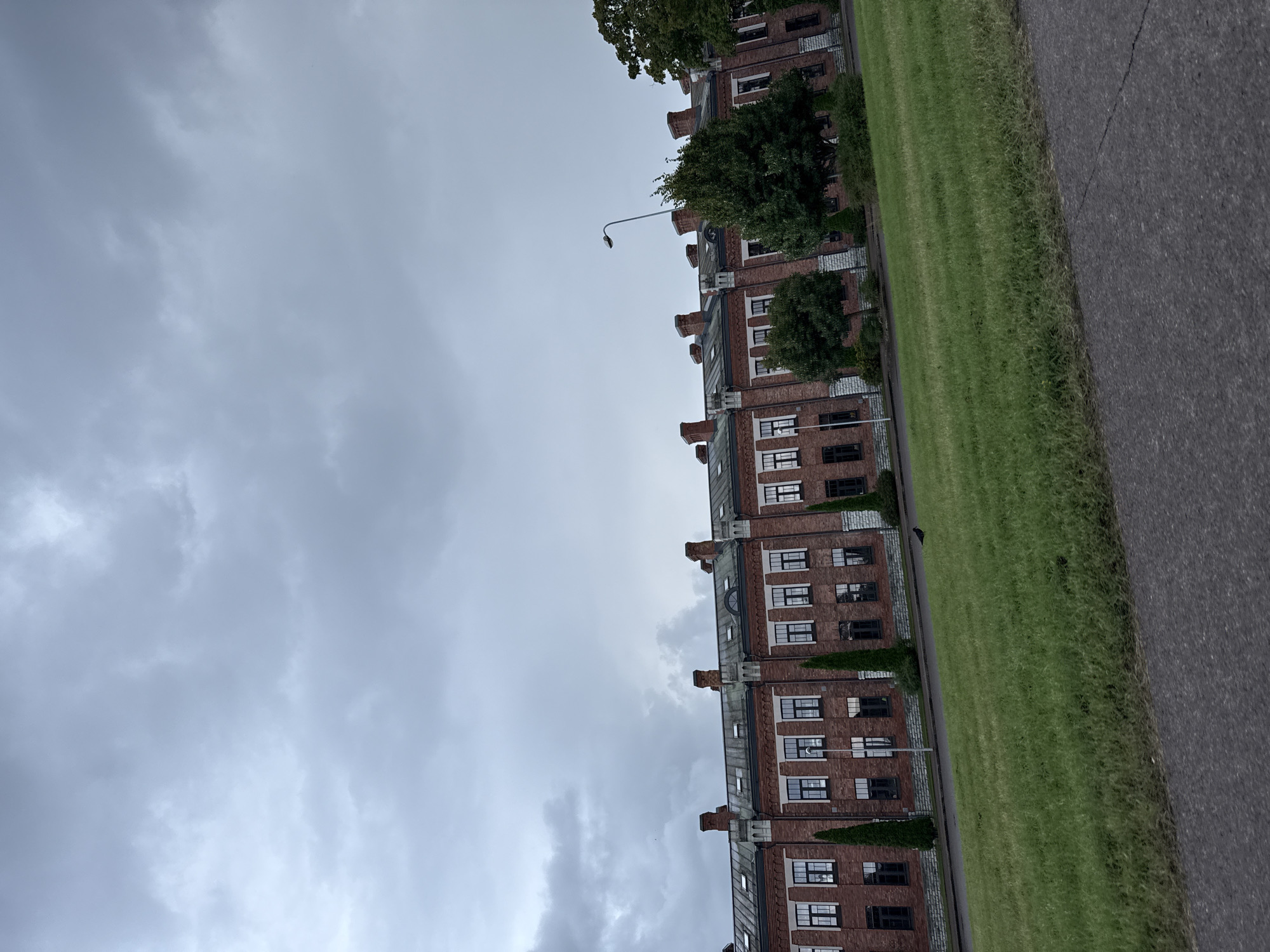 The image depicts a long, multi-story brick building with a dark, overcast sky. The building features numerous windows, many of which appear to be dark or covered. There are several chimney stacks visible on the roof. In front of the building, there is a well-maintained green lawn with some trees and manicured bushes. A street lamp is visible in front of the building. The foreground shows a portion of an asphalt road. A single dark bird is present on the grass in front of the building. The architecture of the building suggests it might be a former industrial or institutional structure. Based on the architectural style and historical context of Tallinn, this building is likely part of the Seaplane Harbour (Lennusadam) complex or a similar historical waterfront development. The cloudy sky indicates a cool or potentially rainy day. There are no people visible in the image.