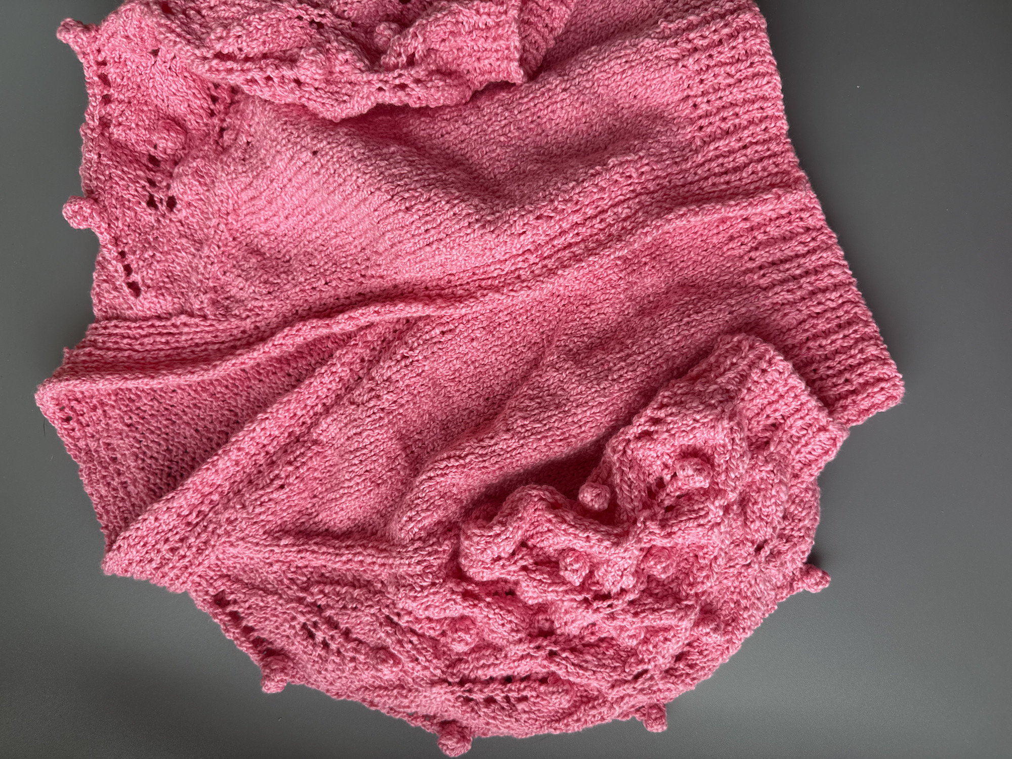 A pink, knitted cardigan is laid out flat on a neutral gray surface. The cardigan features intricate stitch patterns, including what appear to be bobble stitches on the sleeves and along the edges of the neckline, creating a textured and decorative effect. The garment is draped in a way that showcases the details of the knitting. There are no people, actions, or interactions visible in the image. The environment is a plain, indoor setting, likely a table or floor. There is no information in the image to suggest a location in Moscow, Russia.