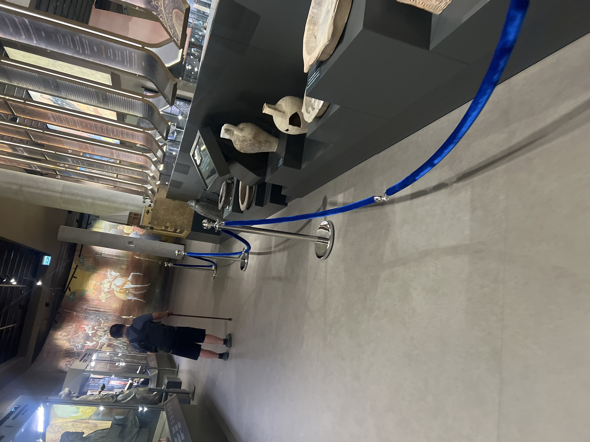 An individual walks in a museum, leaning on a cane for support. They are dressed in a grey shirt and dark shorts. The museum features a large mural depicting historical figures and scenes, and displays of ancient pottery and artifacts. Blue velvet ropes and stanchions are used to guide visitors and protect exhibits. The lighting is dim, with spotlights illuminating key displays. The floor is made of polished concrete.
