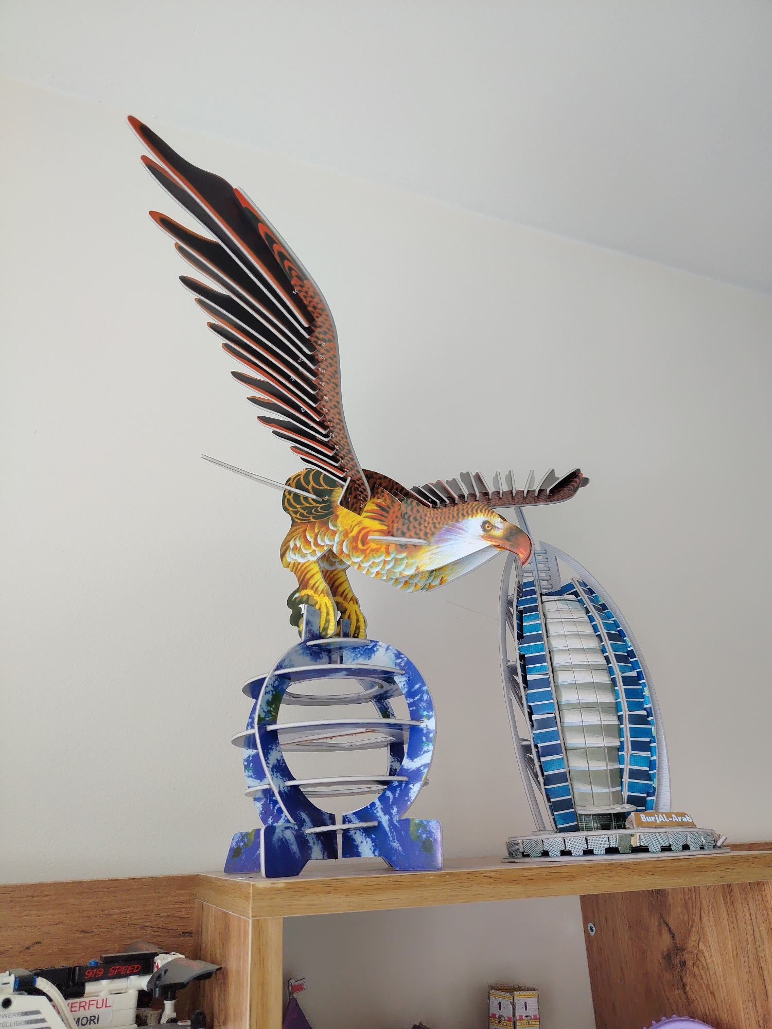This is a close-up, eye-level, medium shot of a shelf displaying several model pieces. In the center of the image, a colorful 3D model of an eagle with outstretched wings dominates the frame, appearing to perch on a blue, globe-like structure. To the right of the eagle is a model of the Burj Al Arab hotel, a sail-shaped skyscraper. The models are made of cardboard and are intricately detailed with printed graphics. The background is a plain, light-colored wall, and the shelf itself is made of wood. The lighting is bright and diffused. The image does not provide any information about Angren, Uzbekistan.