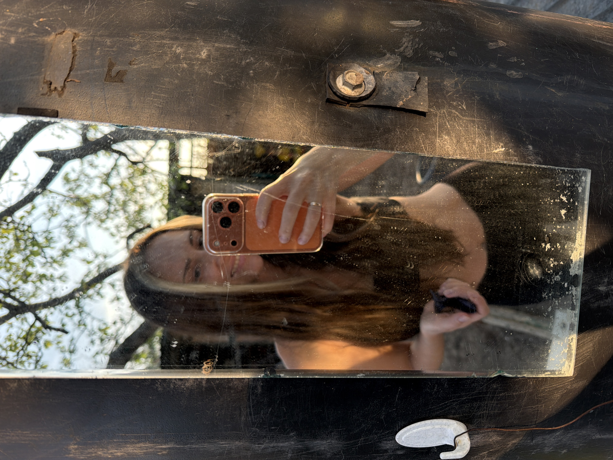 A woman's reflection is captured in a rectangular mirror set against a dark, scratched surface. The woman holds an orange smartphone with three camera lenses, capturing a selfie. In the reflection, trees are visible in the background. A white, hook-shaped object with a thin brown wire attached sits on the dark surface below the mirror. A rusty bolt and metal plate are attached to the upper portion of the dark surface. The location is Kamyshin, Russia.