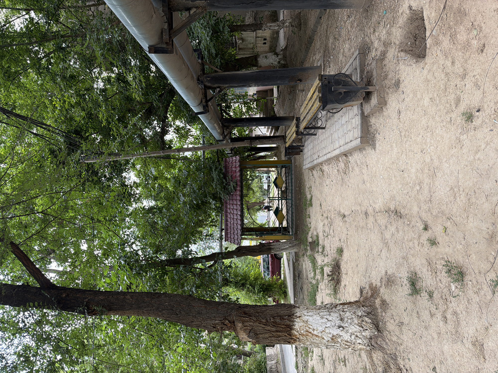 This is a photograph taken outdoors in Shymkent, Kazakhstan. The image is oriented vertically, with the top of the frame showing a view upwards into the canopy of leafy trees and overhead pipes. The scene appears to be a park or public space with a paved walkway leading to a small, covered seating area with a red tiled roof. A park bench with a yellow wooden seat and black metal scrollwork is visible along the walkway, placed on a patterned tile base.
In the background, a red car is partially visible. The ground is a mix of dry soil, scattered twigs, and sparse greenery. The trees are lush and green, suggesting a temperate season, likely spring or summer. The lighting suggests it is daytime, possibly overcast or shaded by the trees, as there are no strong shadows. The overall atmosphere is one of a tranquil, everyday scene in a local park. No people are clearly visible, though a dark shape within the covered seating area might be a person. The image is uncaptioned and contains no visible text.