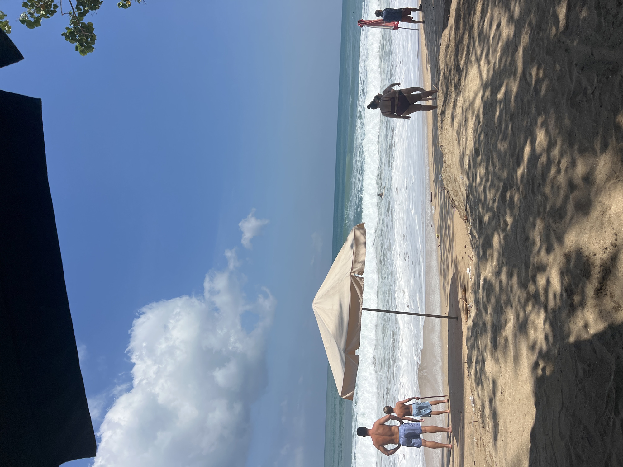 A bright, sunny day unfolds on a sandy beach in Kuta, Indonesia. The turquoise ocean, with its white-capped waves, stretches to the horizon under a clear blue sky dotted with fluffy clouds. A large, beige beach umbrella stands planted in the sand, offering a patch of shade.
In the foreground, two men and a boy stand facing the ocean, their backs to the camera. The man on the left wears blue swimming trunks, and the boy beside him wears similar trunks and holds a stick. Further down the beach, two women in bikinis walk towards the water, their forms silhouetted against the bright backdrop. To the far right, another person stands near a red and white striped umbrella. The sand is textured with shadows cast by unseen trees, suggesting a nearby canopy. A few pieces of driftwood lie scattered on the shore. The overall scene conveys a relaxed, seaside atmosphere.