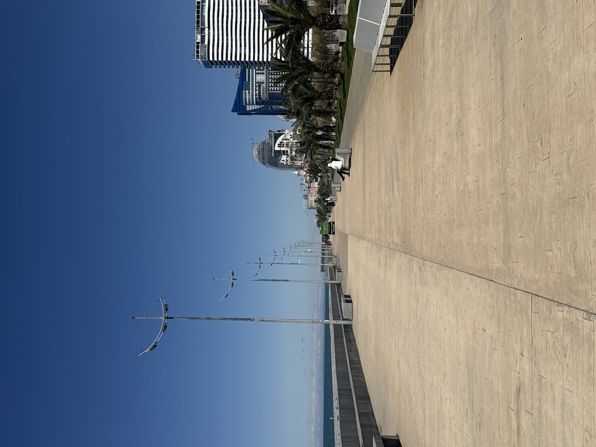 The image depicts a seaside promenade in Batumi, Georgia, on a clear day with a bright blue sky. The walkway is made of light-colored concrete tiles and stretches into the distance. Along the walkway are evenly spaced, tall lampposts with distinctive cross-shaped fixtures at the top. Palm trees and benches line the other side of the promenade. In the background are various modern architectural buildings, including high-rise structures with unique designs. A few people can be seen walking along the promenade. The sea is visible as a narrow strip of blue along the horizon.
