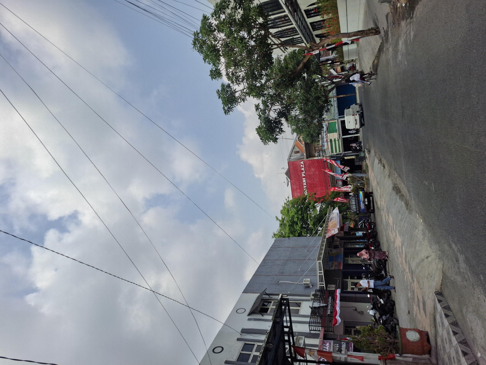 The image shows a street scene in Lamongan, Indonesia. On the left side of the street is a two-story building with a gray facade and windows. The ground floor has shops with awnings and flags. A few motorcycles are parked in front of the shops. Two people are walking on the sidewalk. On the right side of the street is another building with a white facade and arched windows. A large tree with green foliage stands in front of this building. A white van is driving down the street, and a person on a bicycle is also visible. The sky is partly cloudy. The scene appears to be a commercial or mixed-use area.