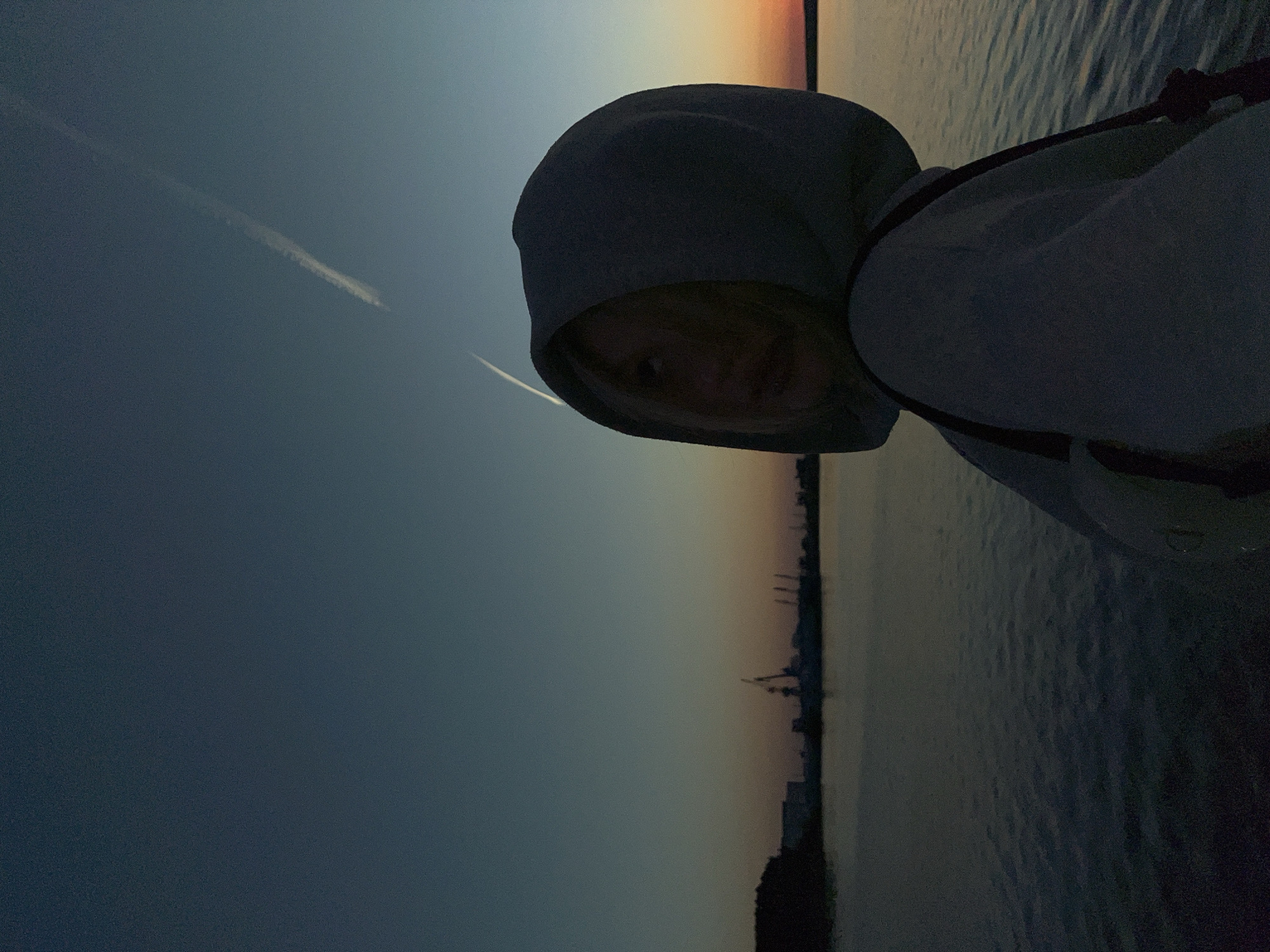 A medium, angled shot depicts a person in a light-colored hooded sweatshirt looking at the camera. The person's face is partially obscured by the hood. In the background, there is water and the silhouette of what appears to be a cityscape during a sunset. In the sky, there is a contrail. Given the prompt, the cityscape is likely Chaikovsky, Russia. The exact location within Chaikovsky is not discernible from this image.