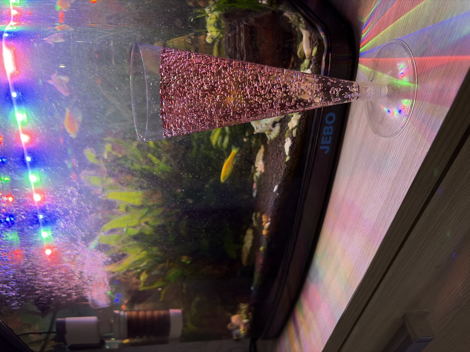 In the city of Central, Russia, a tall, clear plastic glass filled with a pink, bubbly beverage stands on a wooden surface in the foreground. The glass has a conical shape, tapering to a thin stem and a wide, flat circular base. The beverage inside is effervescent, filled with numerous small bubbles that catch the light. Behind the glass, an aquarium is visible. The aquarium contains a dark substrate at the bottom, with various green aquatic plants, and at least one small yellow fish swimming. The aquarium is illuminated from above by a string of colorful LED lights, casting streaks of red, green, and blue light onto the wooden surface and the glass. The aquarium itself has a black trim with the word "JEBO" subtly visible on the front. The overall scene suggests a dimly lit, possibly celebratory or relaxed setting, with the aquarium lights and the bubbly drink creating a festive atmosphere.