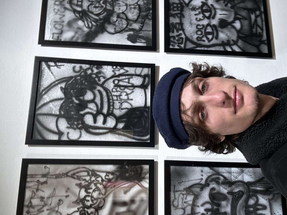 A fair-skinned person with light brown hair and a dark blue beret stands in front of a white wall featuring framed black and white artwork. The artwork appears to be graphic and features spray-painted designs. The person is wearing a dark-colored, textured jacket and is positioned to the right of the framed artwork. The image is slightly angled, giving a candid, selfie-like feel.