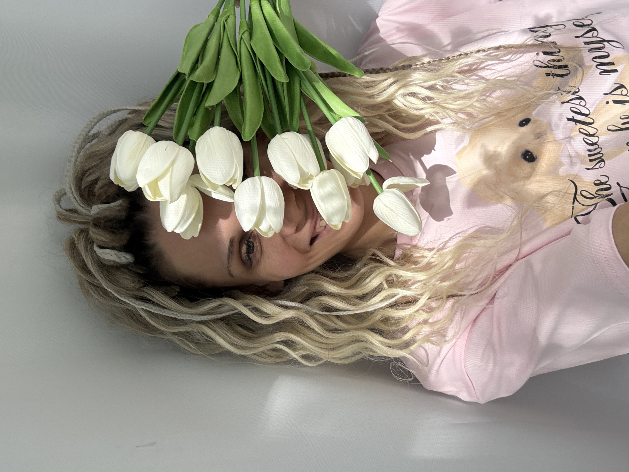 A woman with blonde braided hair lies in a white space. She is wearing a pink t-shirt with an image of a bear and text on it. She is holding a bouquet of white tulips with green stems and leaves above her head, near her face. The location is inferred as Rusanovo, Russia.