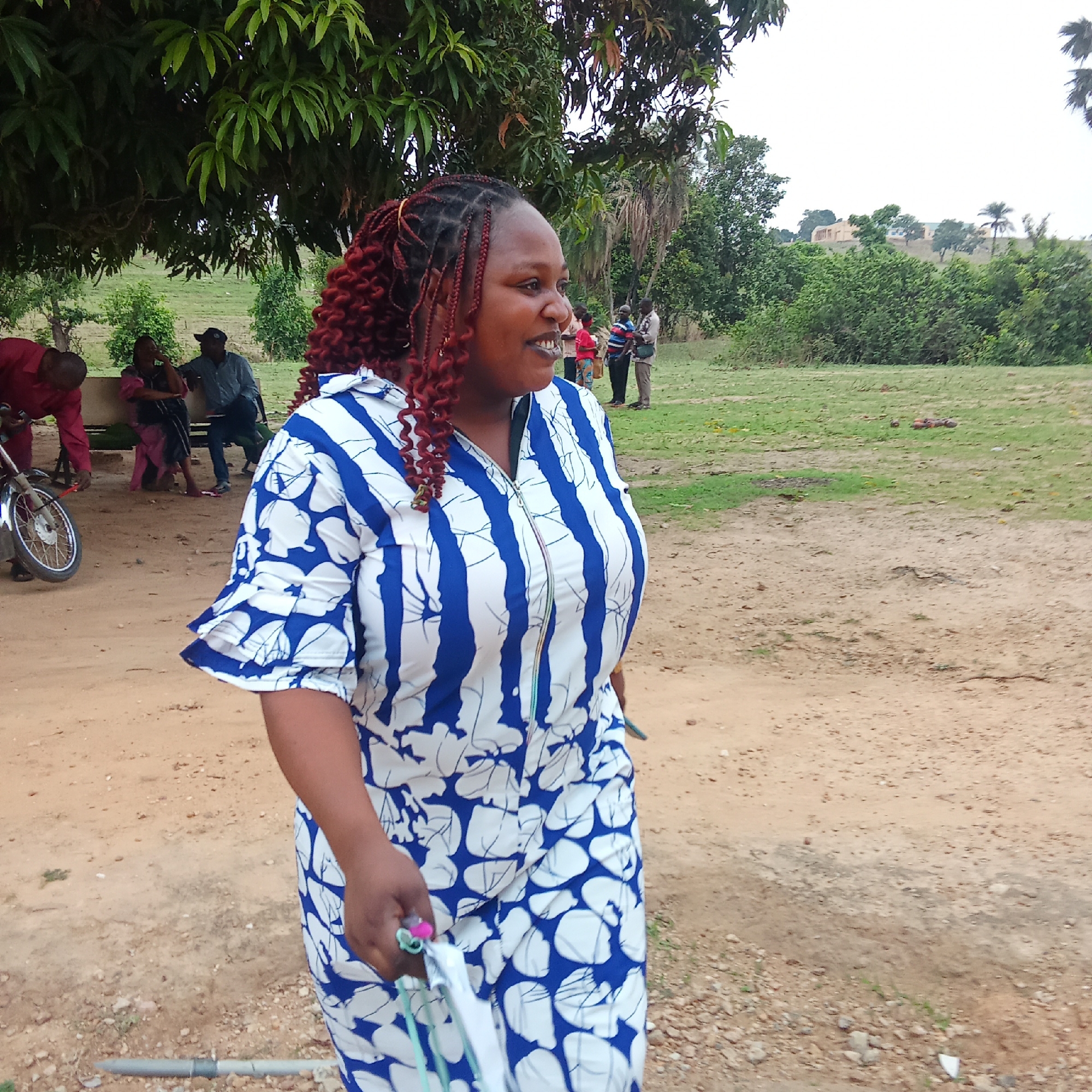 The image shows an African woman in Akwanga, Nigeria, walking on a dirt road in a rural area. She is wearing a blue and white patterned dress with a zipper front and red-tinged braided hair. In the background, there is a group of people under a tree, some sitting and others standing. A motorbike is parked nearby. The environment consists of a dirt road, grassy areas, trees, and vegetation. A building is visible in the distance.