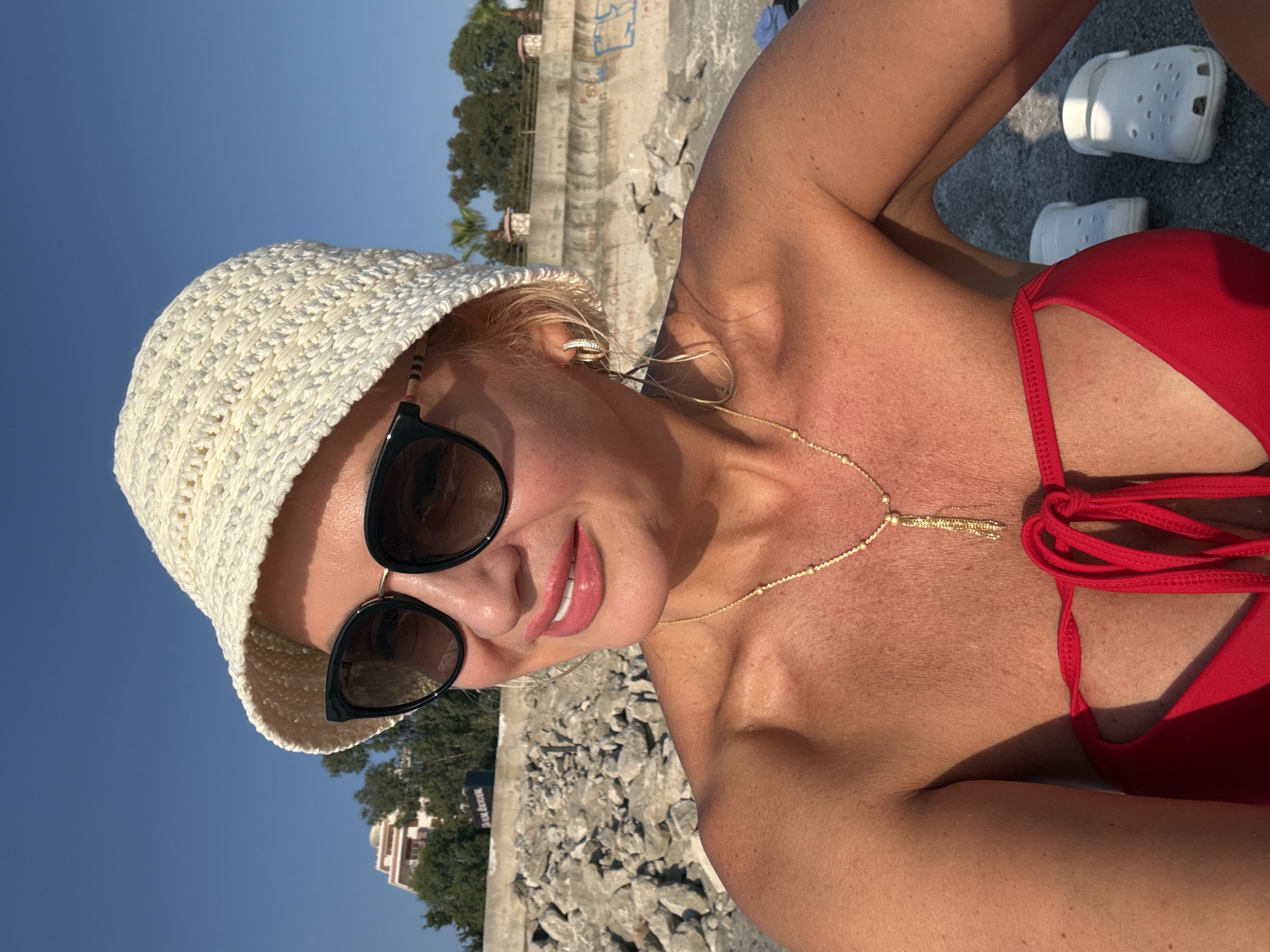 A blonde woman, wearing a red bikini, a white crocheted bucket hat, and sunglasses, poses for a selfie on a rocky beach in Alanya, Turkey. She has a gold necklace with a tassel pendant and small hoop earrings. In the background, there is a concrete sea wall with graffiti, trees, and buildings. Two white Crocs shoes are visible on the ground. The sky is clear and blue, and the sun is shining.