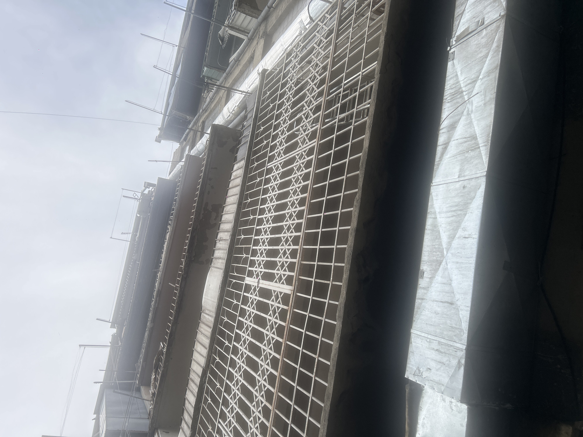 An upward-angle, full-shot photograph shows the exterior of a multi-story building, likely in Tbilisi, Georgia, under an overcast sky. The building's facade is visible, featuring multiple balconies protected by white, collapsible metal grates. The grates have a diamond pattern and appear to be old and somewhat weathered. Below the balconies, large, rectangular metal ducts run horizontally. These ducts are also dirty and show signs of wear. Electrical wires and conduit pipes are attached to the building's exterior, running vertically alongside the walls. The overall impression is one of a dated and somewhat neglected urban environment. The image is somewhat hazy, suggesting either atmospheric conditions like fog or a low-quality camera sensor. There are no people visible in the image.