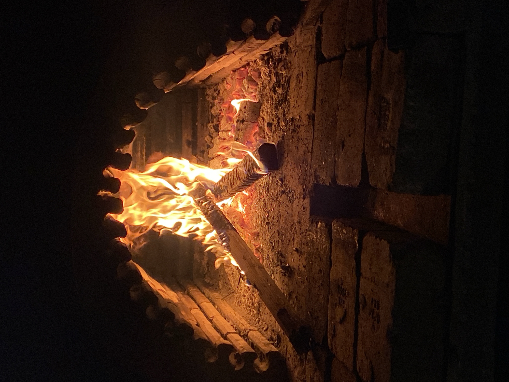 An indoor fireplace is lit, consuming logs within a stone structure with a decorative, notched front. The scene is predominantly dark, highlighting the bright orange and yellow flames. The fireplace walls are constructed of rough-hewn stones. Konstantinovo, Bulgaria, is not discernible from the image.