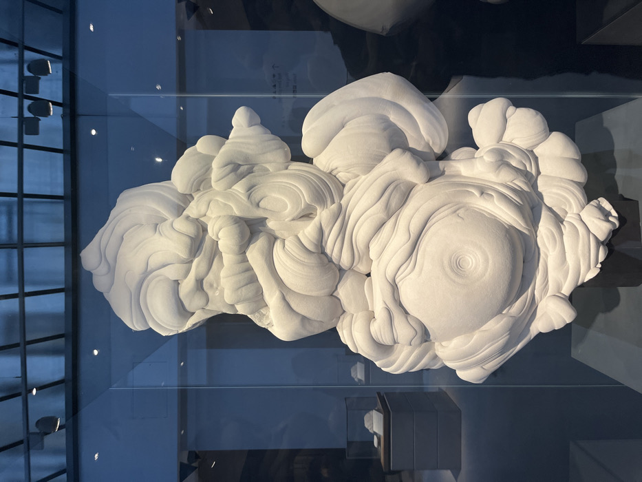 A large, intricate, abstract sculpture is displayed indoors within a modern exhibition space, likely a contemporary art gallery or museum, in the city of London, United Kingdom. The sculpture, primarily white or off-white, is composed of highly textured, undulating, and layered organic forms, resembling solidified foam, clouds, or geological formations. Its surface features numerous rounded protuberances and flowing patterns, including a prominent concentric circular design on a lower, central lobe. The artwork stands on a dark, indistinct pedestal and is viewed through a clear glass panel, which reflects elements of the ceiling and distant background.
The immediate environment behind the sculpture is dimly lit with a blueish hue, revealing additional, smaller white sculptural pieces displayed in glass cases on grey pedestals further back. The ceiling above features a dark-framed grid structure, possibly a skylight, with several recessed spotlights. Reflected in the glass panel, particularly in the upper right quadrant, is a white directional sign with an upward arrow and text that includes East Asian characters (likely Japanese). No individuals are visible in the scene. The overall ambiance is sterile and curated. The location is consistent with a major contemporary arts venue such as the Barbican Centre.