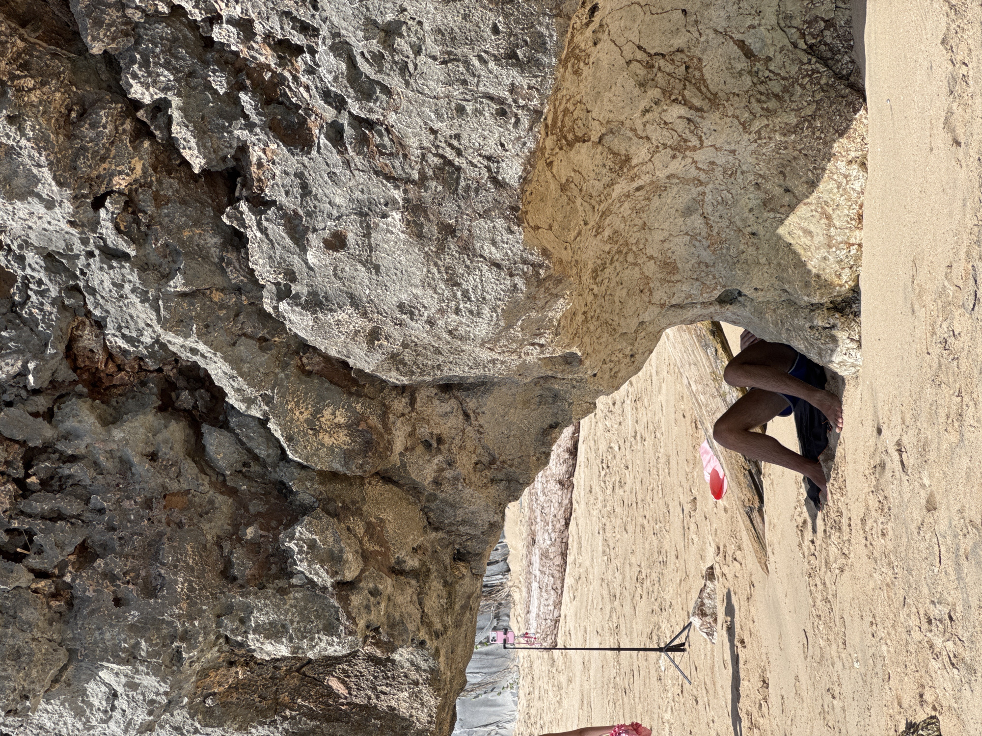 The image shows a view of a rocky cliff face in Kuta Selatan, Indonesia, likely taken from a beach or sandy area. The composition is angled, presenting the cliff as a dominant, vertical structure. The rock formations are varied in color and texture, with darker, rougher sections juxtaposed against smoother, lighter-colored areas. A small opening or cave is visible at the base of the cliff, partially obscuring a person's legs. The person is wearing blue swimwear and is barefoot. Next to them, there are what appear to be a pair of pink sandals. Further along the sandy ground, there's a camera tripod set up. The scene is lit by natural sunlight.